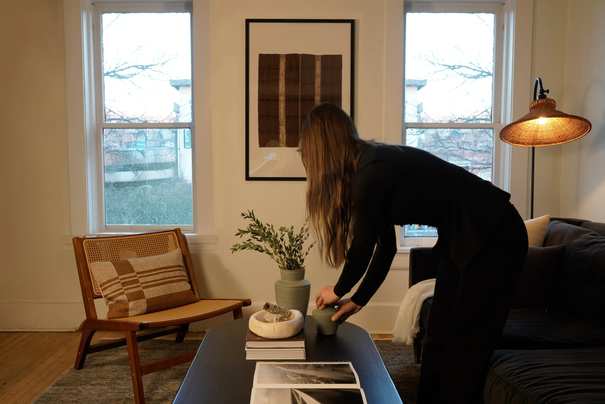 A person with long hair, dressed in black, arranging objects on a coffee table in a living room with two windows, a framed artwork, a potted plant, and a floor lamp.