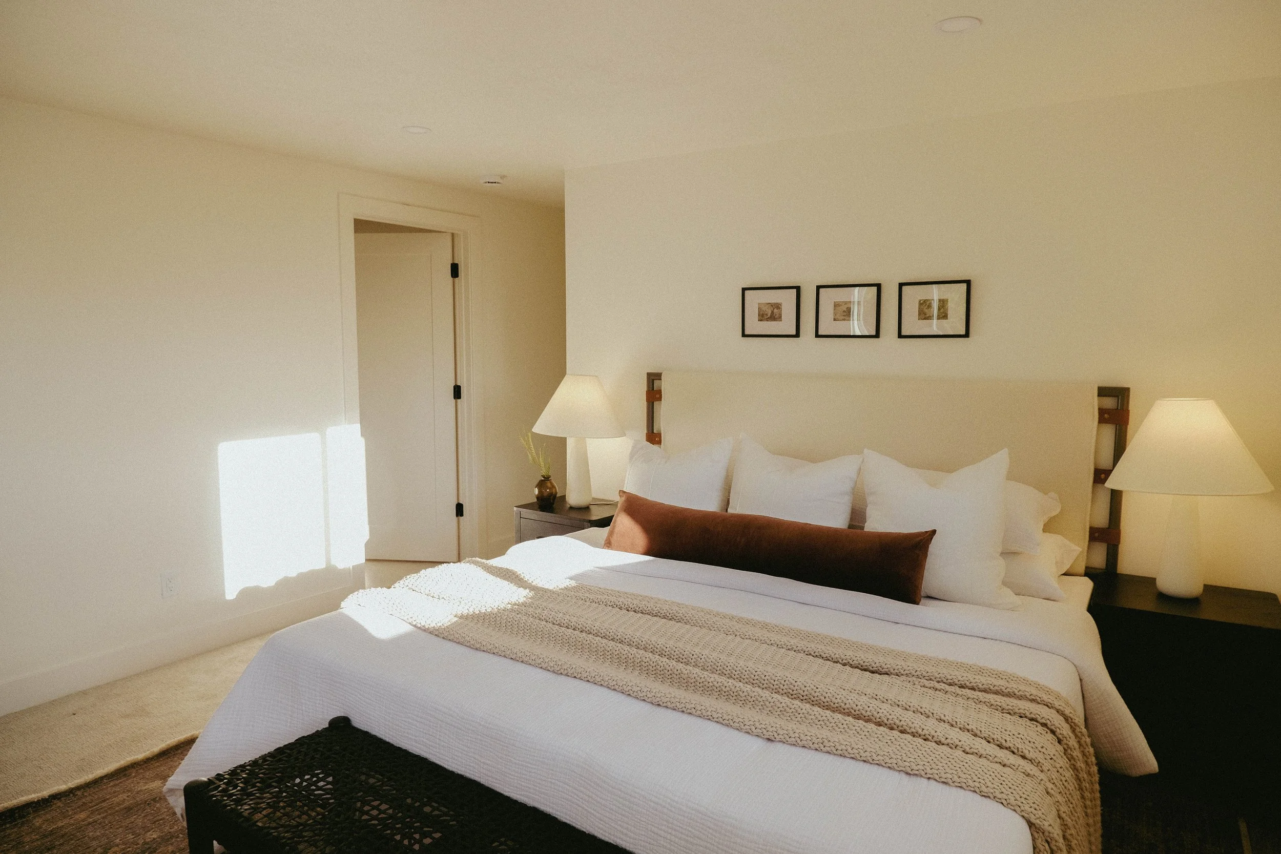 A neatly made bed with a white comforter, brown pillow, and beige throw blanket in a cozy bedroom with beige walls, framed artwork, a nightstand with a lamp, and sunlight streaming in.
