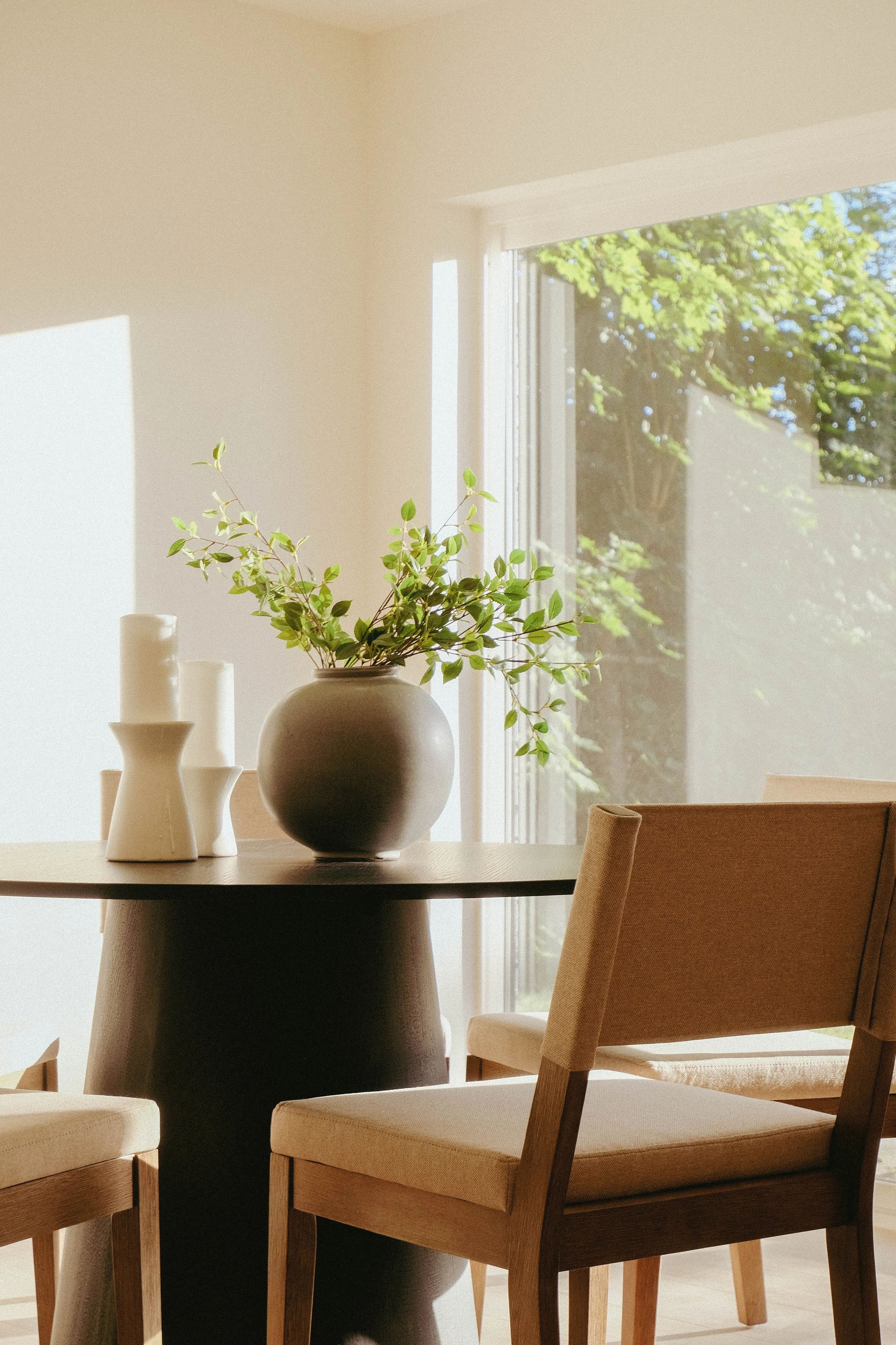 A dining room with a round black table, white chairs, a large round white vase with green leaves, and a window with sunlight streaming in, showing trees outside.