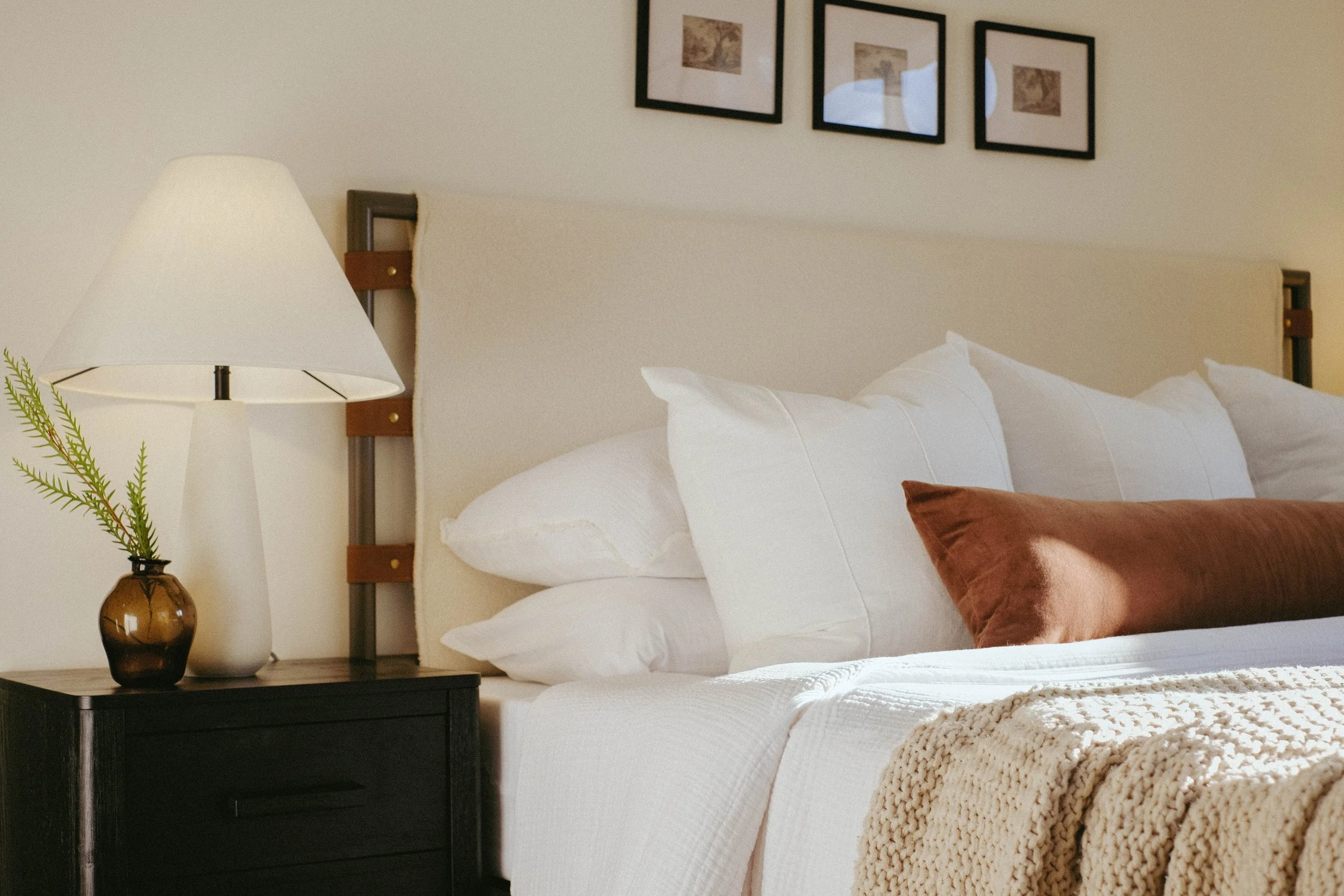 A bedroom with a neatly made bed featuring white bedding and pillows, a brown pillow, and a chunky knit beige blanket. Next to the bed is a black nightstand with a small brown vase holding green fern leaves. A white lampshade sits on a lamp on the ni