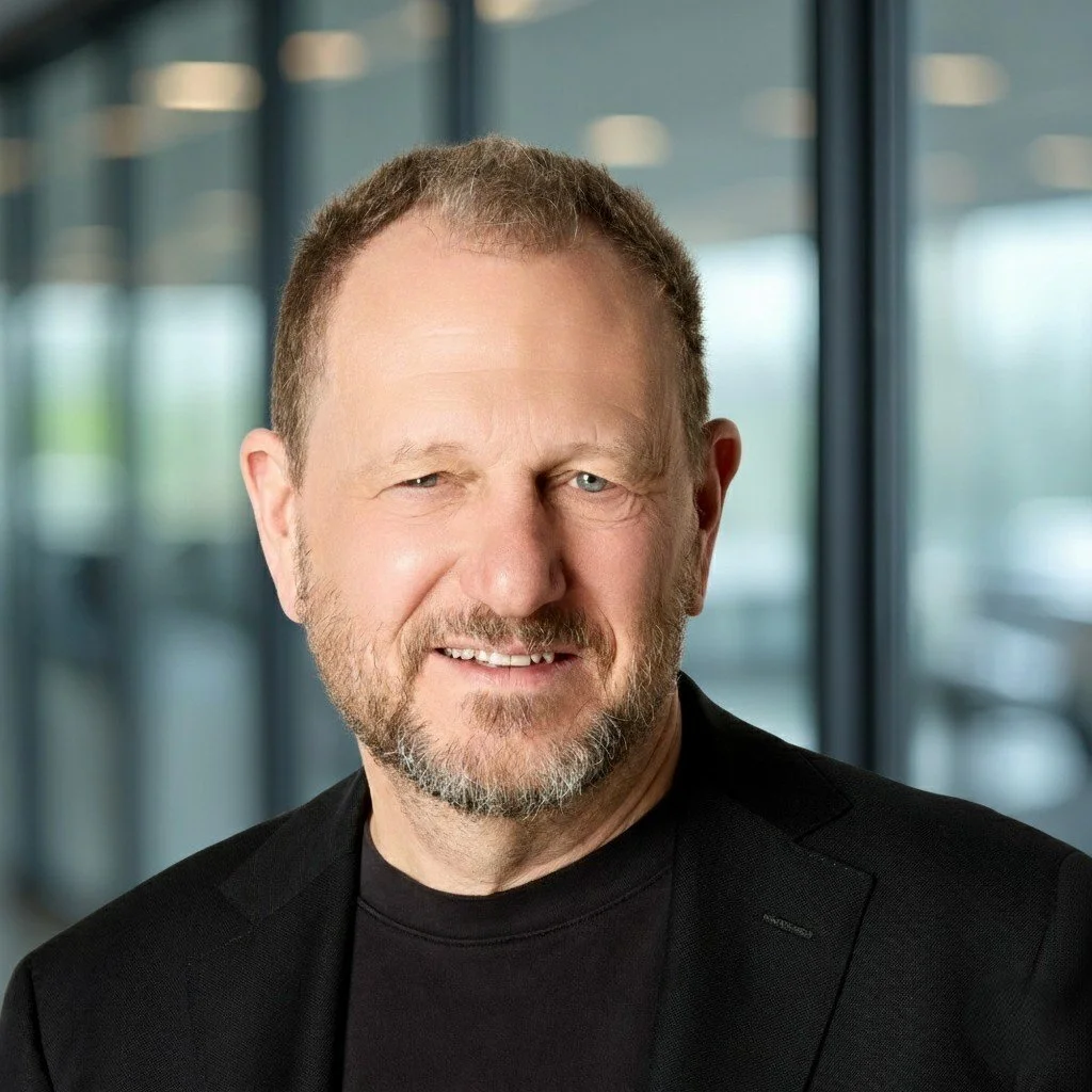 A professional headshot of a man wearing a dark suit, white striped shirt, and black tie, smiling against a blue background.
