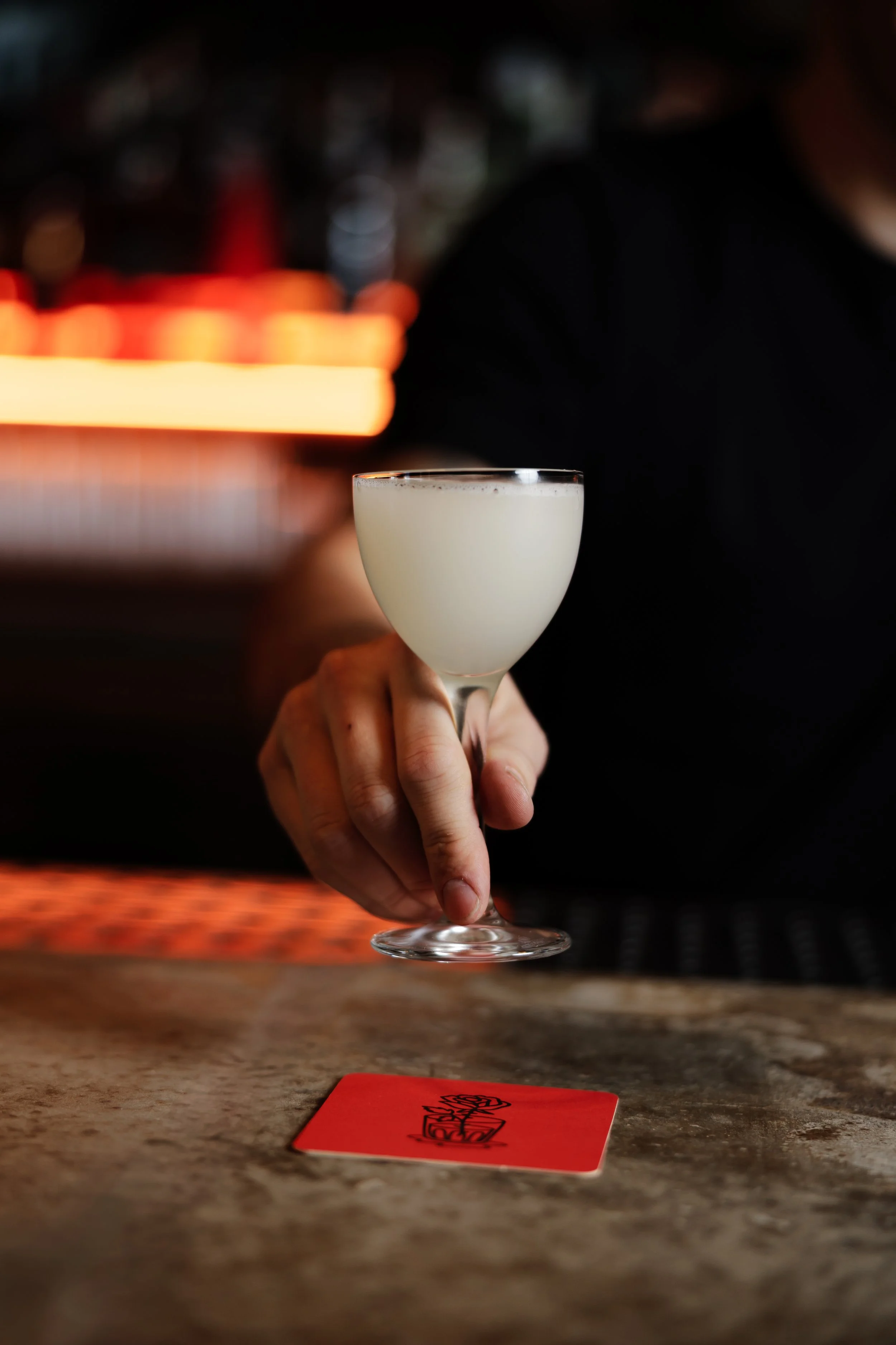 A bartender or server holding a cocktail glass filled with a pale, creamy drink, positioned over a bar counter with a red card on it, in a dimly lit bar or restaurant setting.