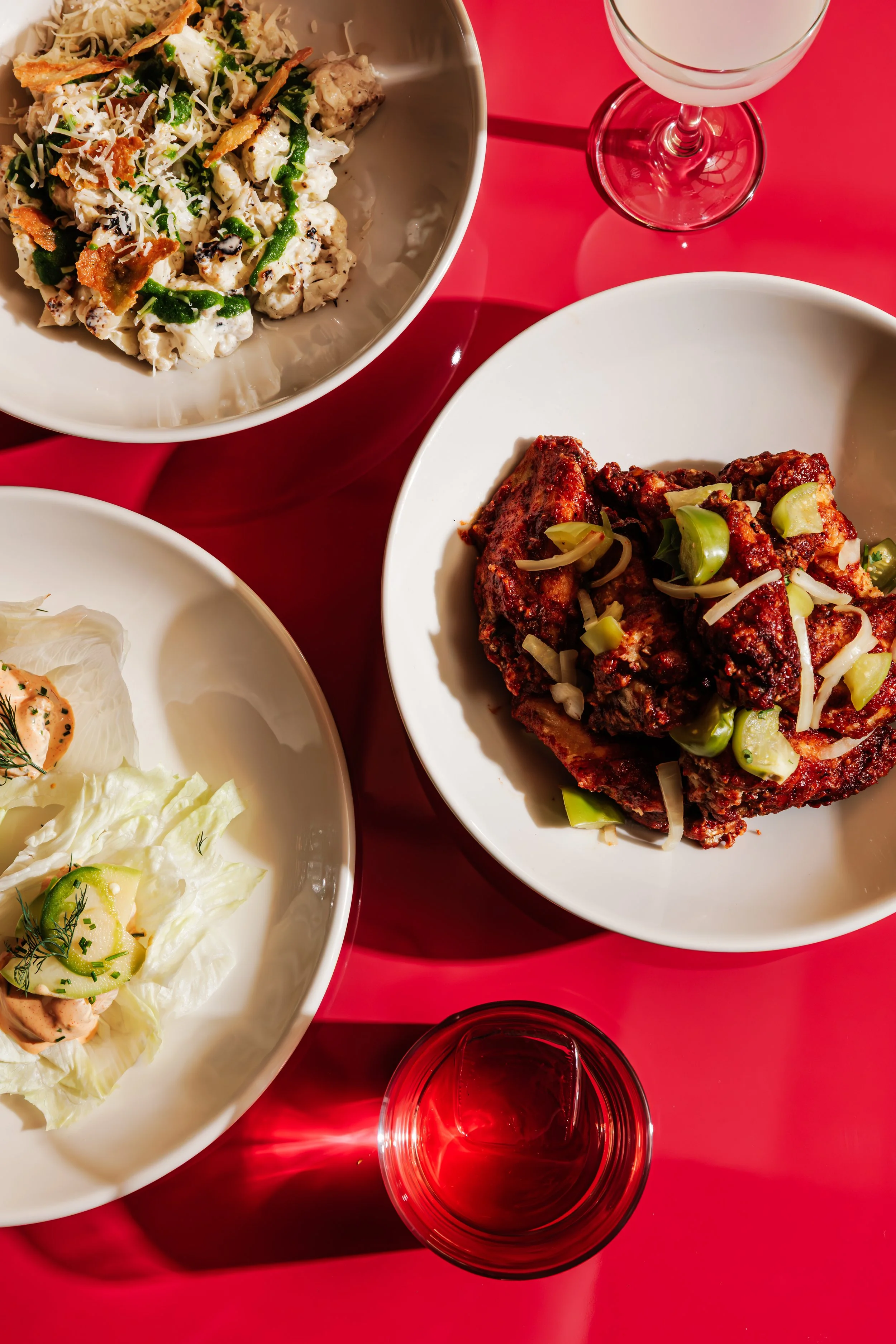 Three plates of food on a red table with two glasses of wine, including a bowl of creamy chicken salad with broccoli and cheese, a plate of spicy ribs with chopped scallions, and an appetizer with lettuce, smoked salmon, and cucumber slices.