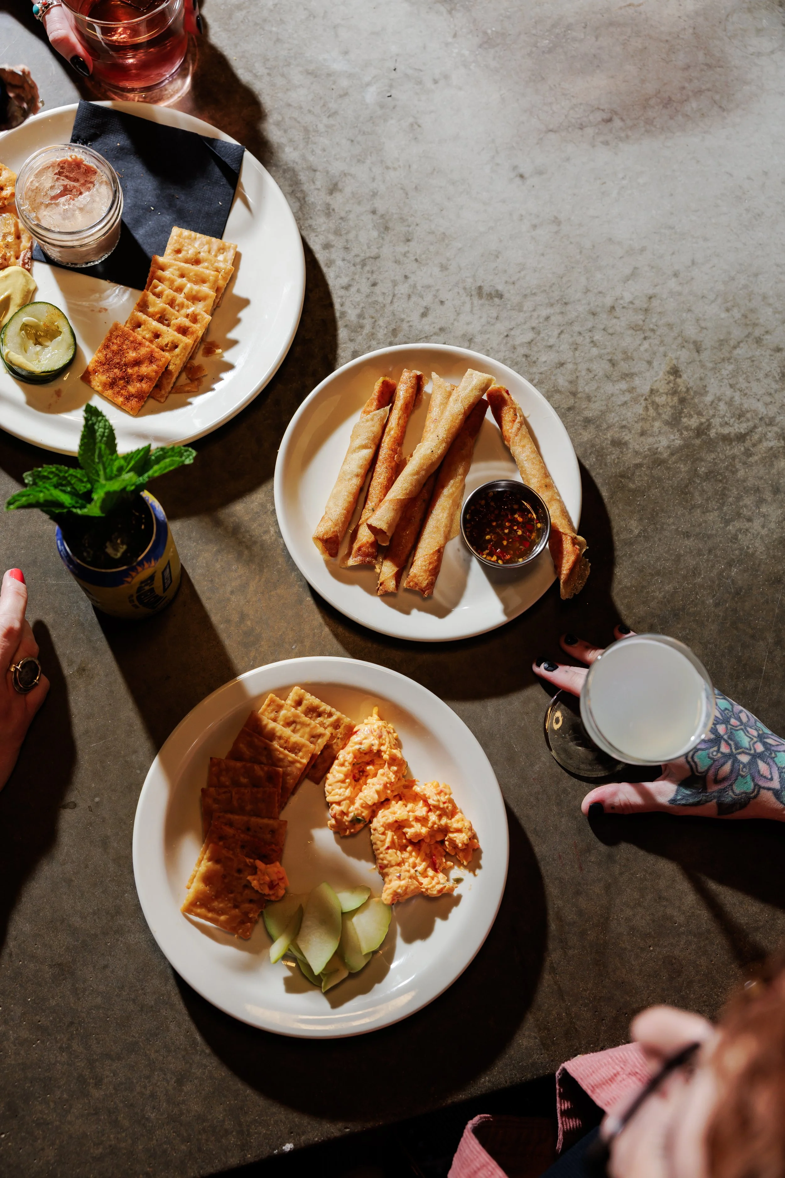 Several white plates with snacks, a glass of drink with a white beverage, a small jar of sauce, and a small plant in a purple container on a gray table, viewed from above.