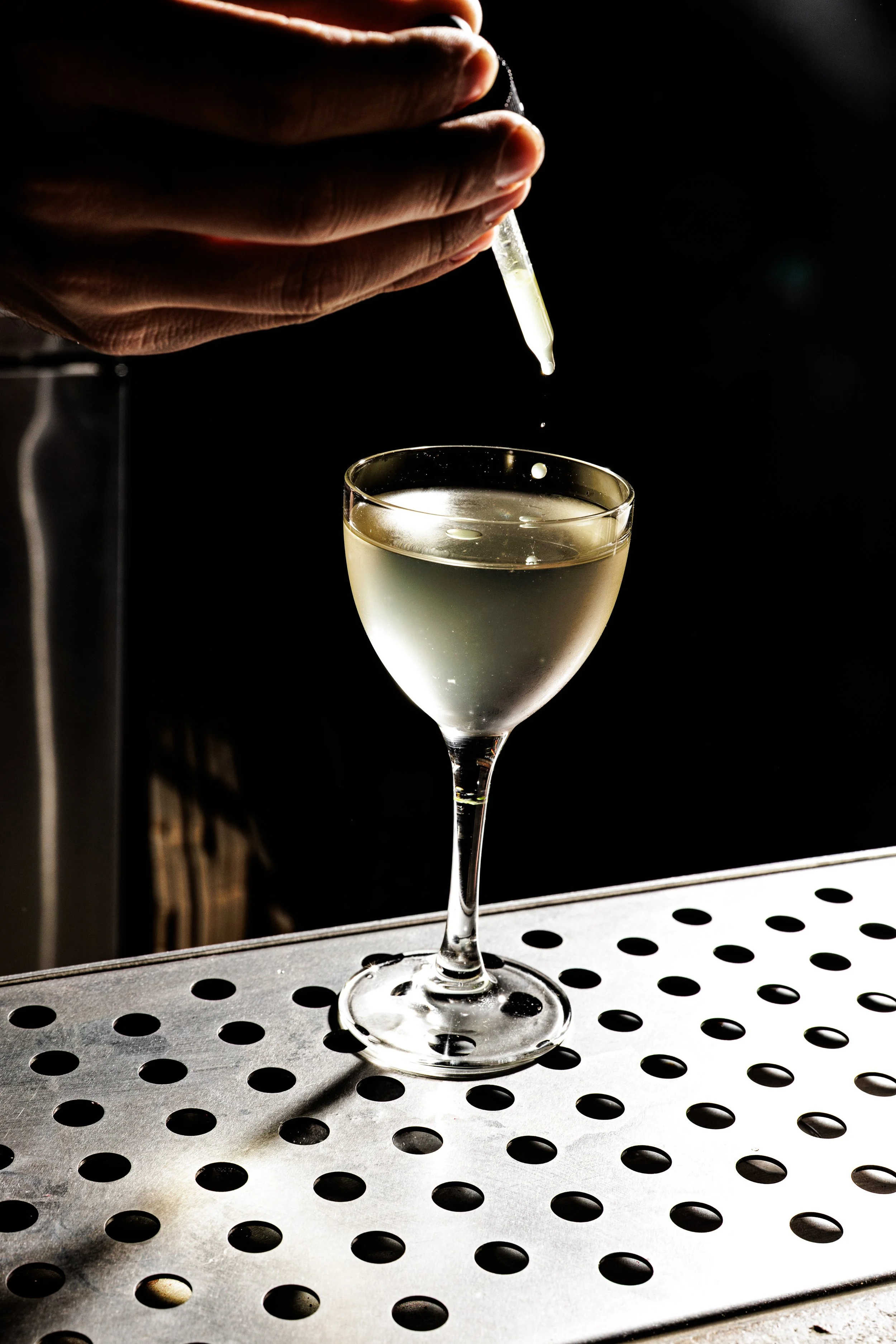 A bartender drops a liquid from a dropper into a glass of white wine on a bar counter.