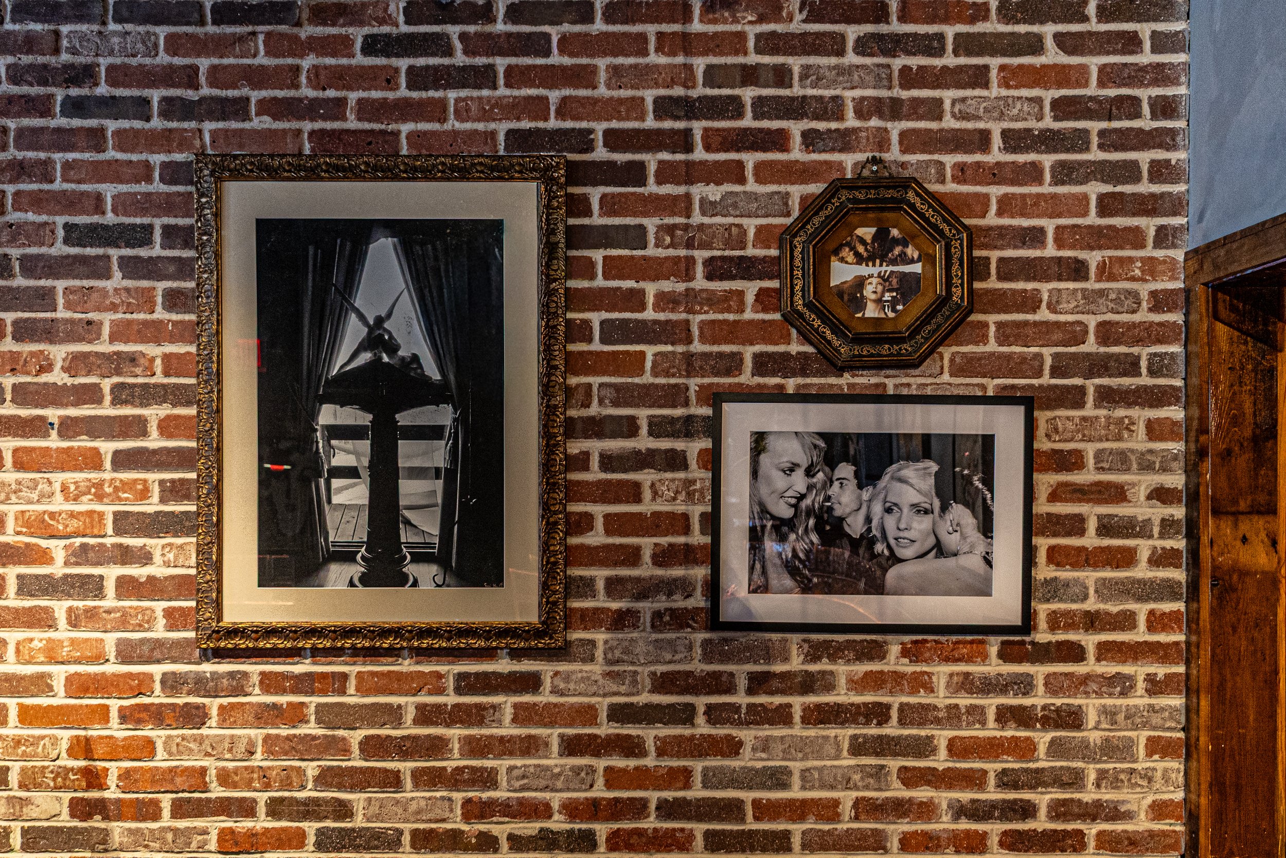 Love's Alibi Nashville interior design. A brick wall decorated with three framed black-and-white photographs. The photo on the left features a woman sitting at a table with a lamp, curtains, and window behind her. The middle photo shows two women in 