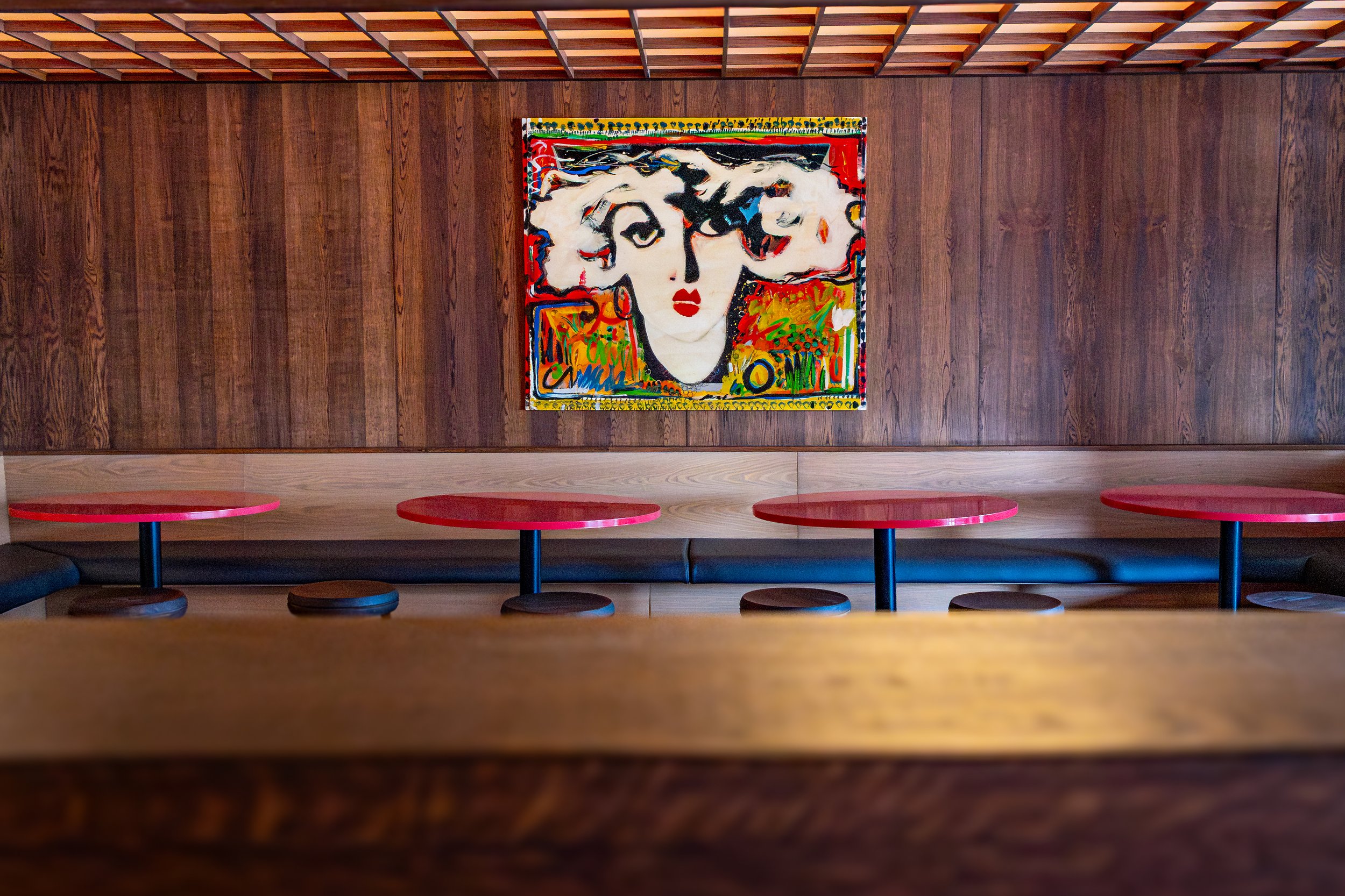 A colorful abstract portrait painting of a woman's face hangs on a dark wooden wall in a room with red circular tables and black booth seating.