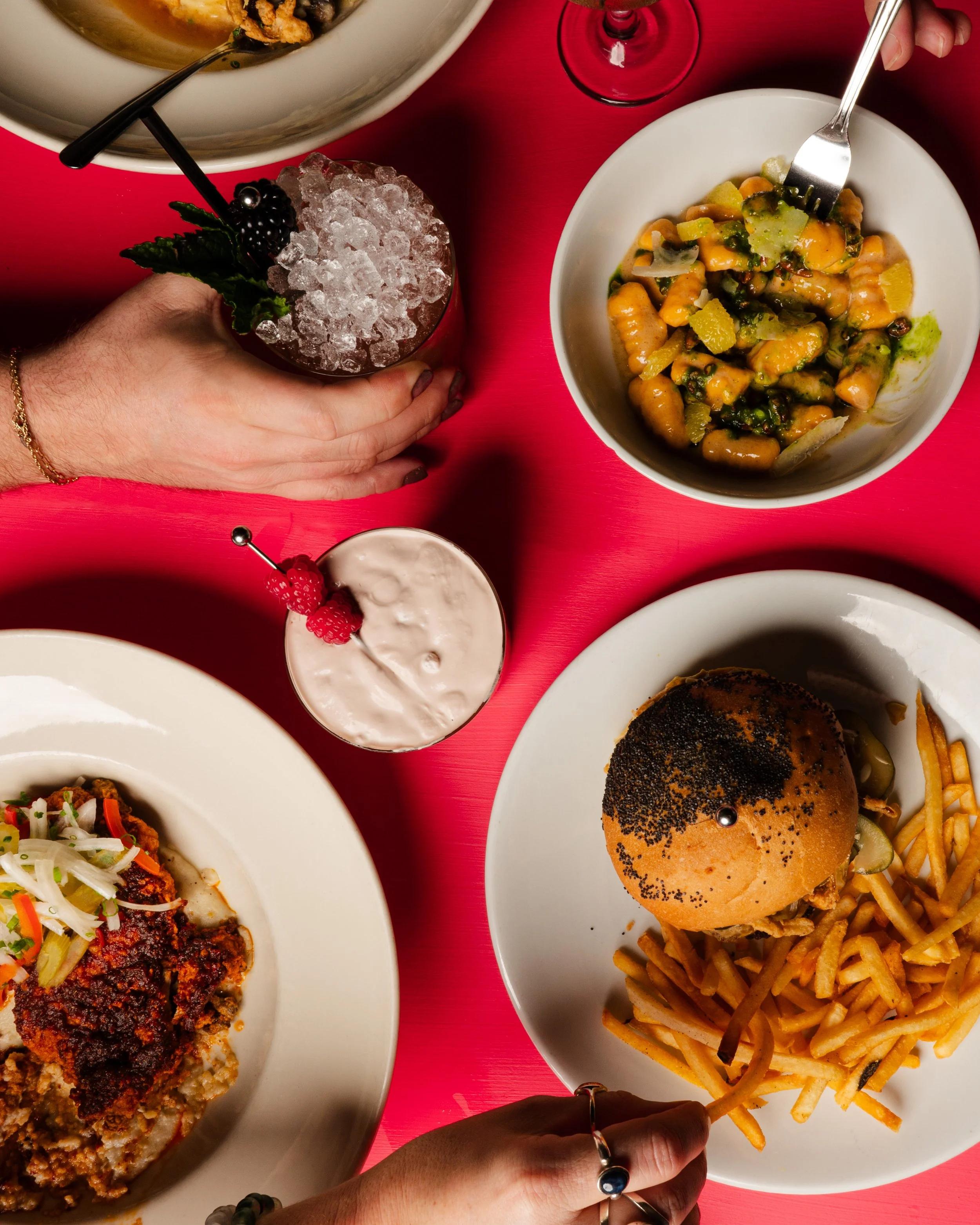 Top-down view of a vibrant table with various dishes including a burger with fries, pasta salad, a plate of meatloaf with gravy and vegetables, and drinks with garnishes, set on a red table.