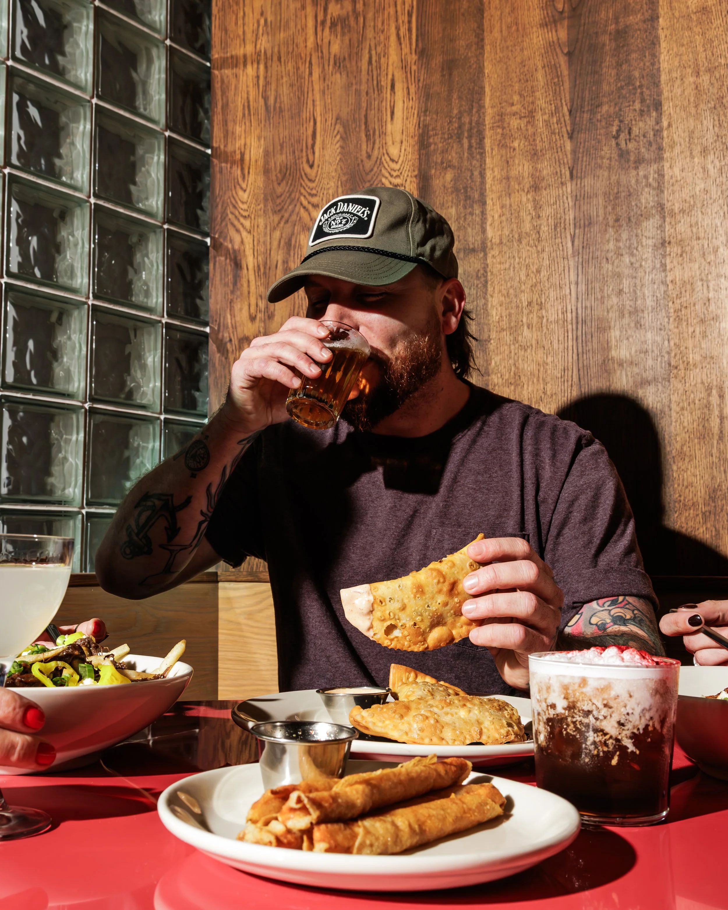 A man with a beard wearing a Jack Daniel's hat and a dark T-shirt, sitting at a table in a restaurant, drinking beer from a glass and holding a piece of fried food, with various dishes including spring rolls, a salad, and a dark cola in front of him.