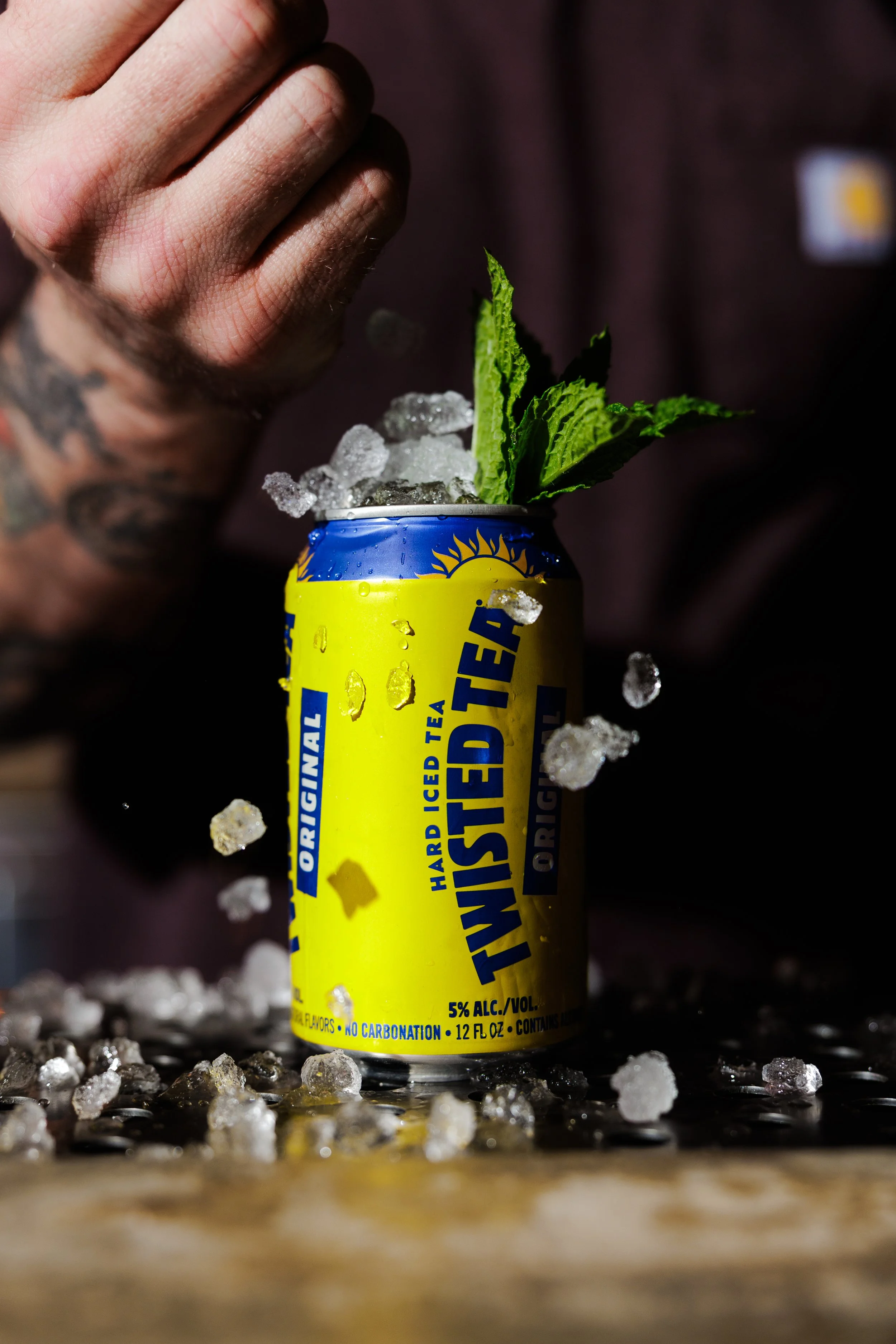 A can of yellow and blue Twist Tea with ice and a mint sprig inside, with scattered ice on a dark surface.