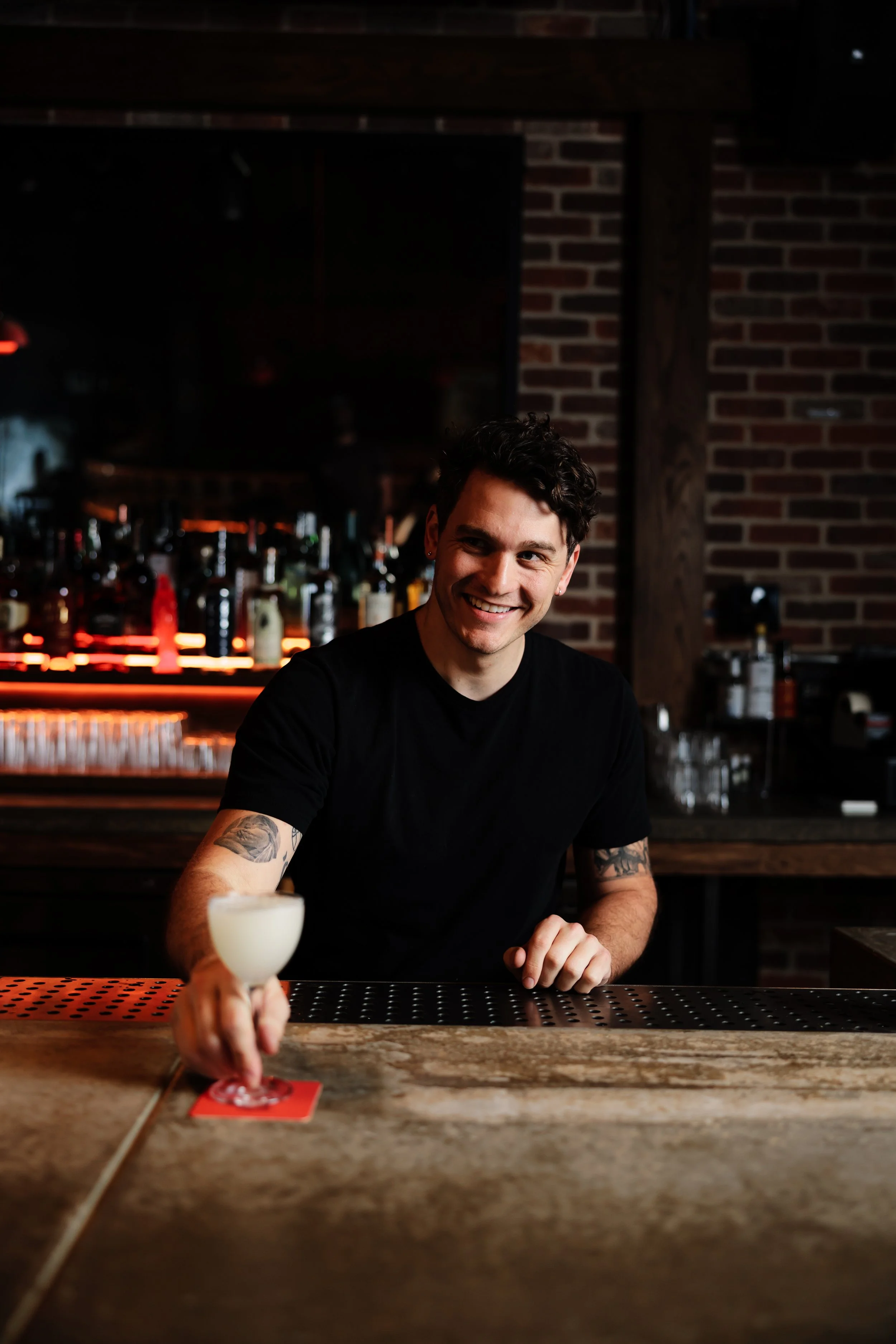 A smiling man with dark curly hair, wearing a black t-shirt, sitting at a bar, reaching for a cocktail with a white frothy top, in a dimly lit bar with bottles in the background.