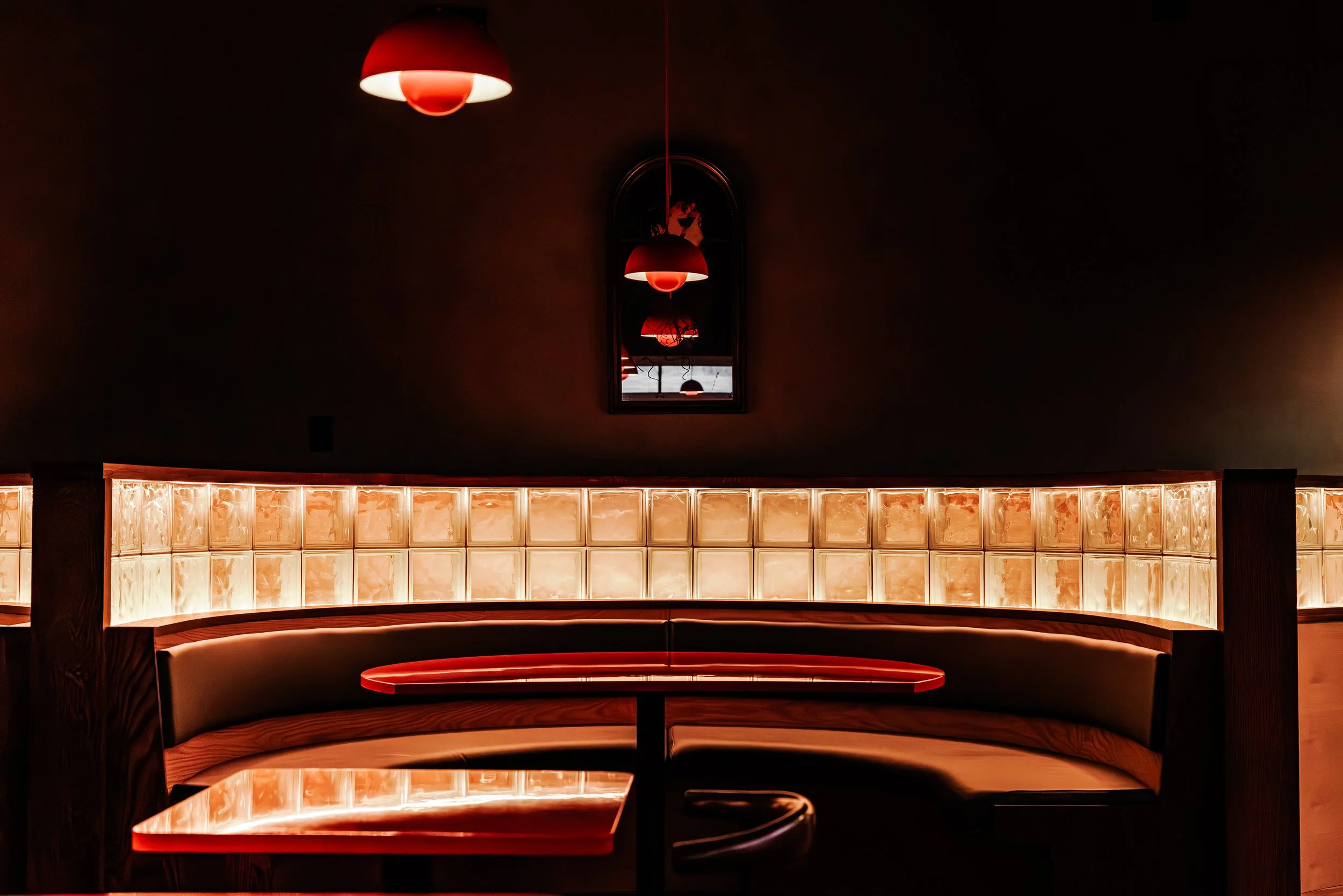 Empty booth seat with a red illuminated table in a dimly lit restaurant or bar with red pendant lamps and a mirror on the dark wall.
