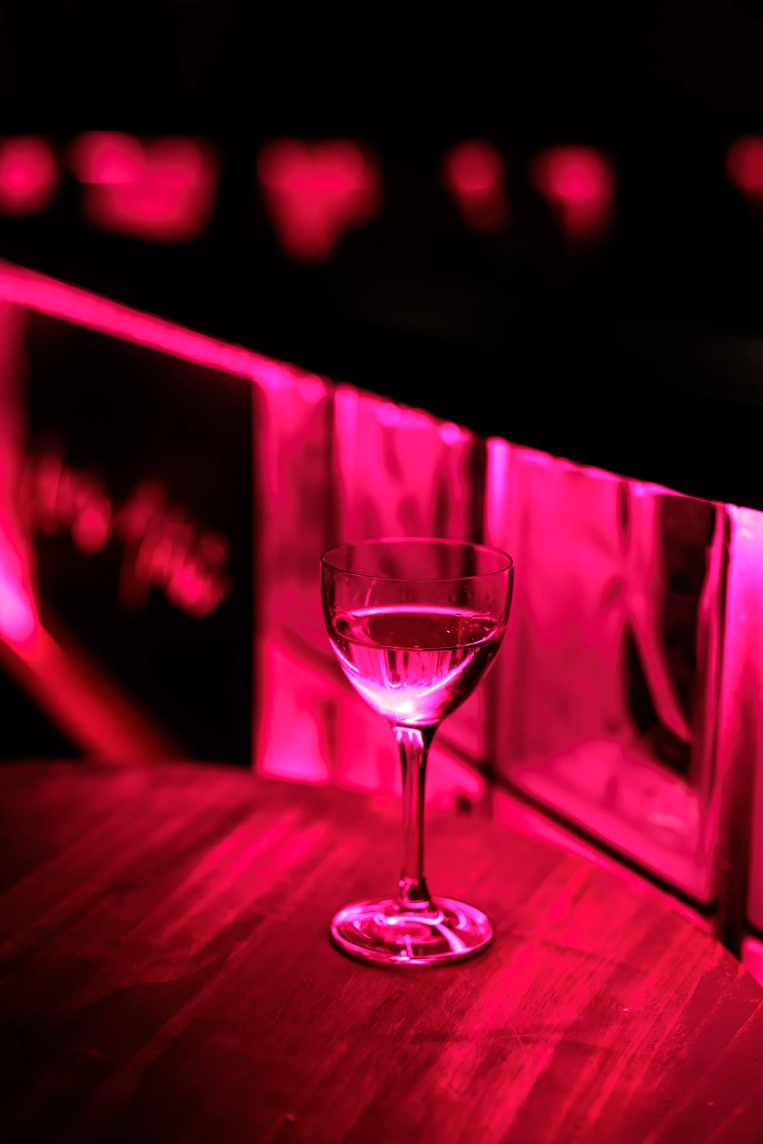 A glass of water on a wooden table illuminated by pink neon lights, with a blurred background featuring pink and black elements.
