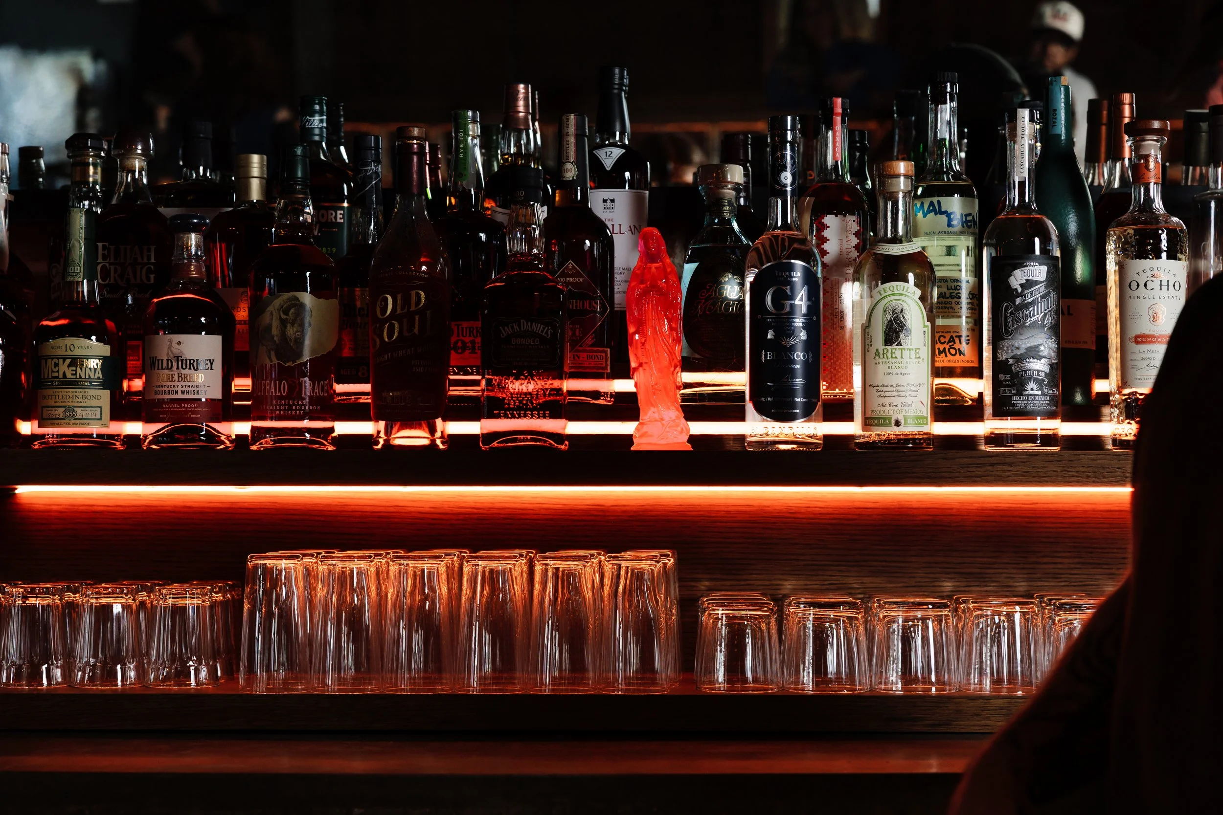 Various bottles of alcohol on a bar shelf illuminated by red lighting, with empty glasses underneath.