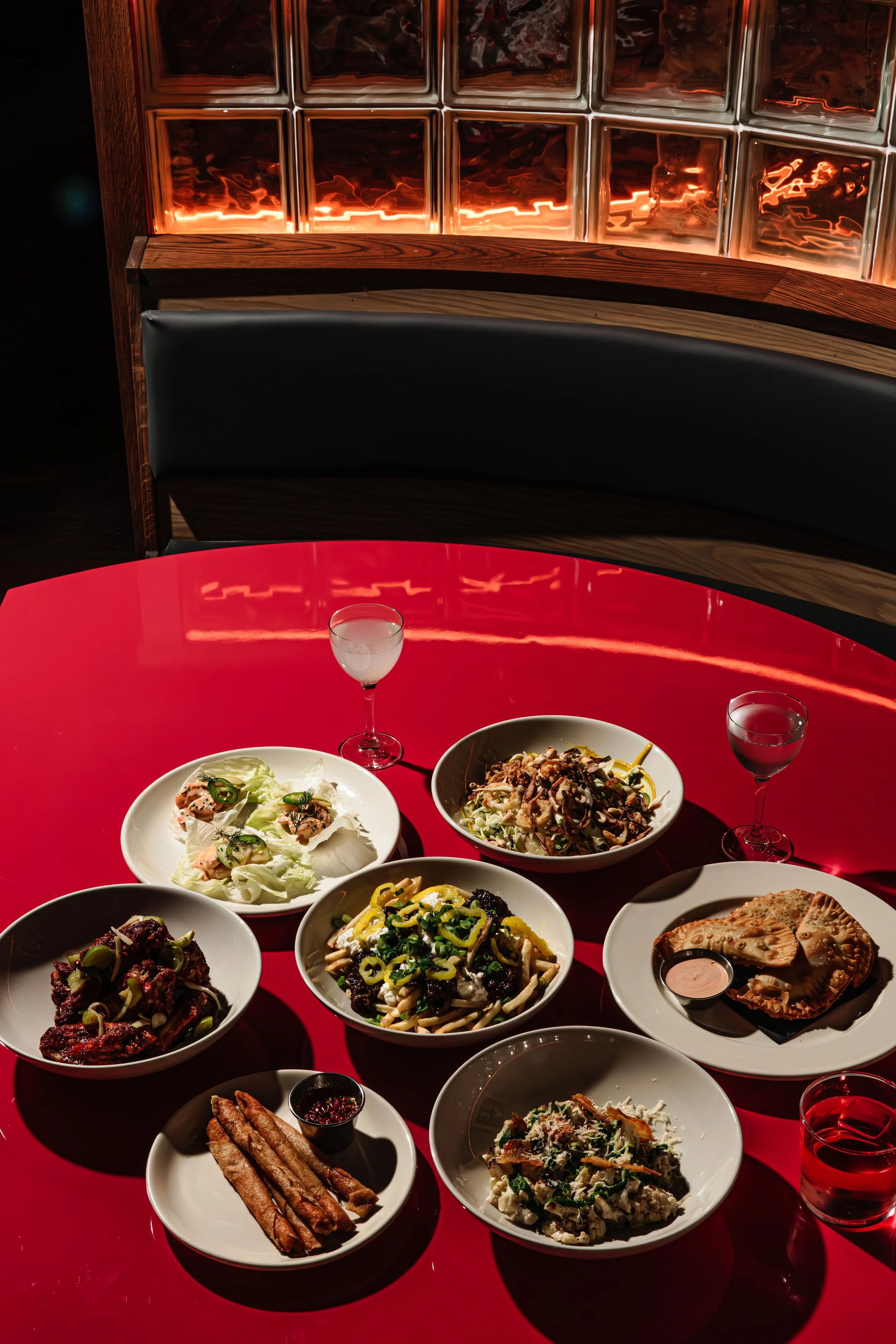 A red table with several plates of food, including salads, pasta, and meat dishes, along with glasses of water in a dimly lit restaurant with a glow of neon light reflection on the wall.