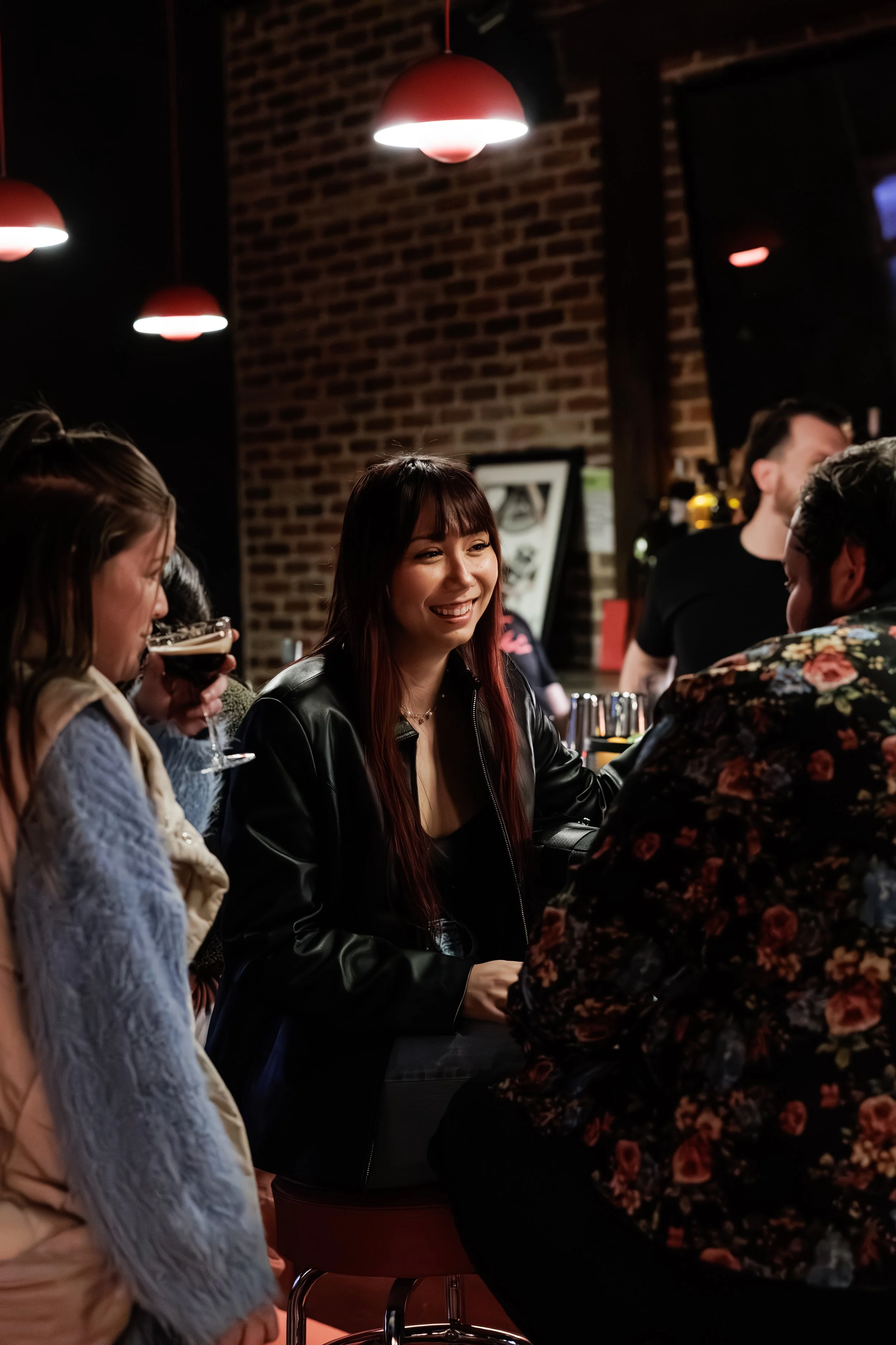 A group of friends socializing in a dimly lit bar with brick walls. They are chatting and drinking, with a woman in the center smiling.