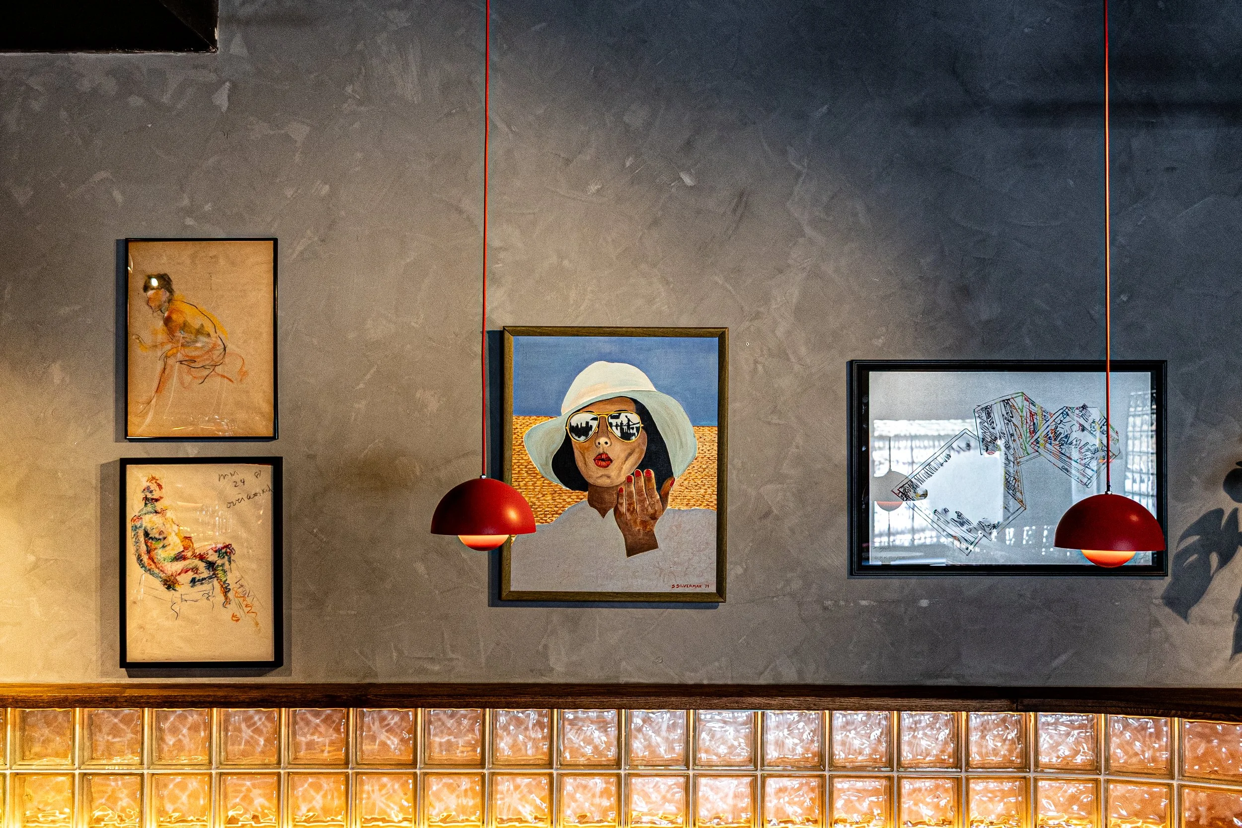 Interior art gallery wall with framed artwork at Love's Alibi Nashville. Including a portrait of a woman wearing sunglasses and a hat, flanked by red pendant lights and a glass-block wall below.