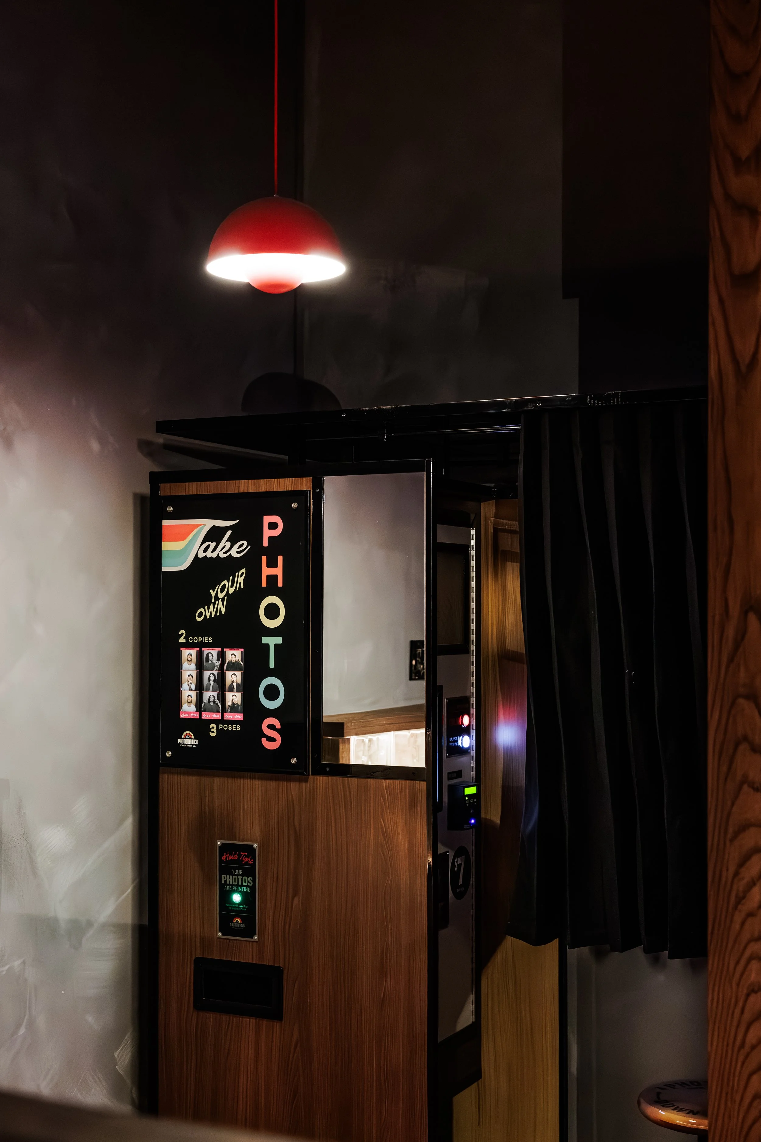 A photo booth with a sign that says 'Take your own photos' and offers two copies and three poses, inside a room with a red hanging lamp and dark walls.