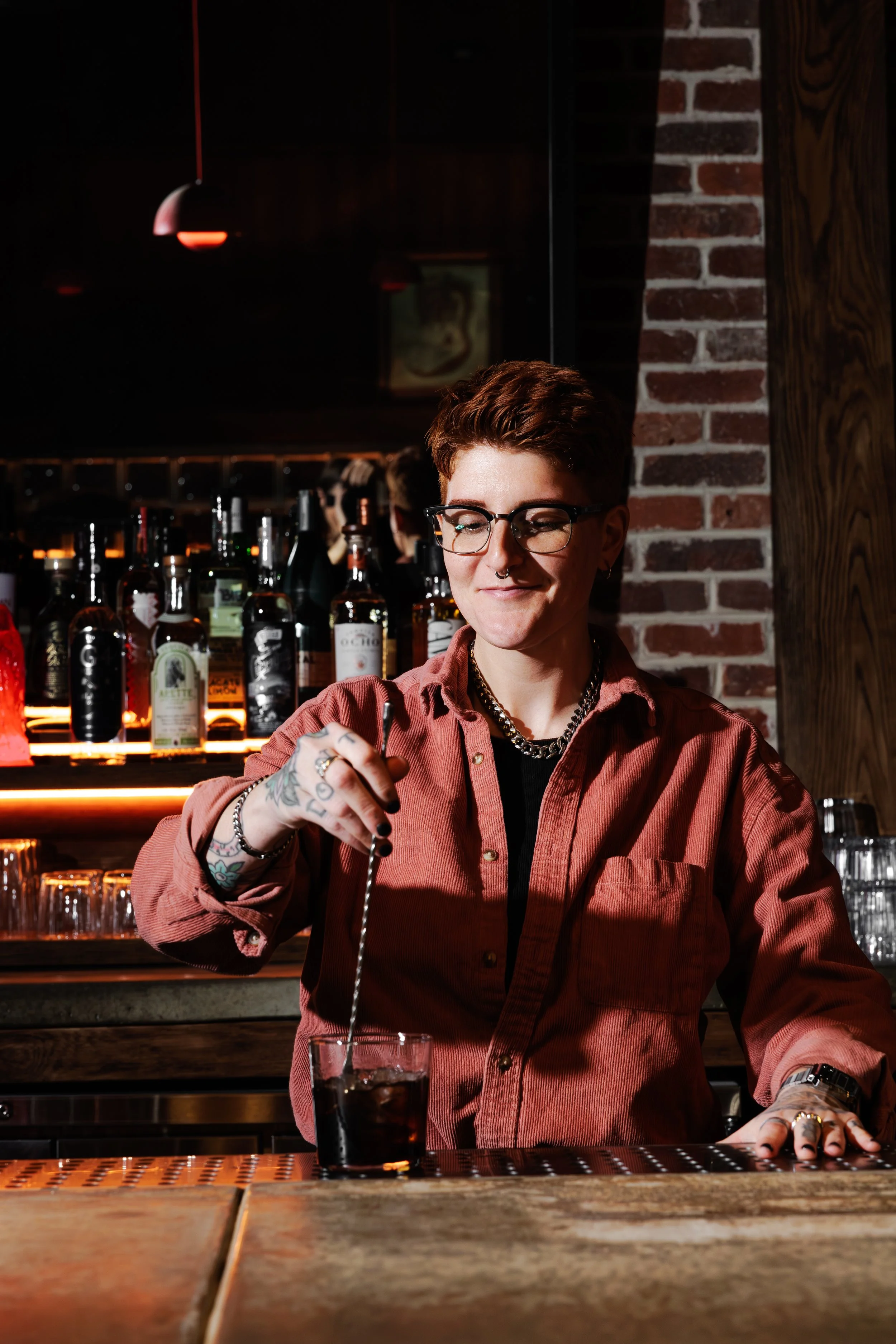 A bartender with short hair, glasses, tattoos, and jewelry is stirring a dark drink at the bar in a dimly lit bar with bottles of liquor in the background.