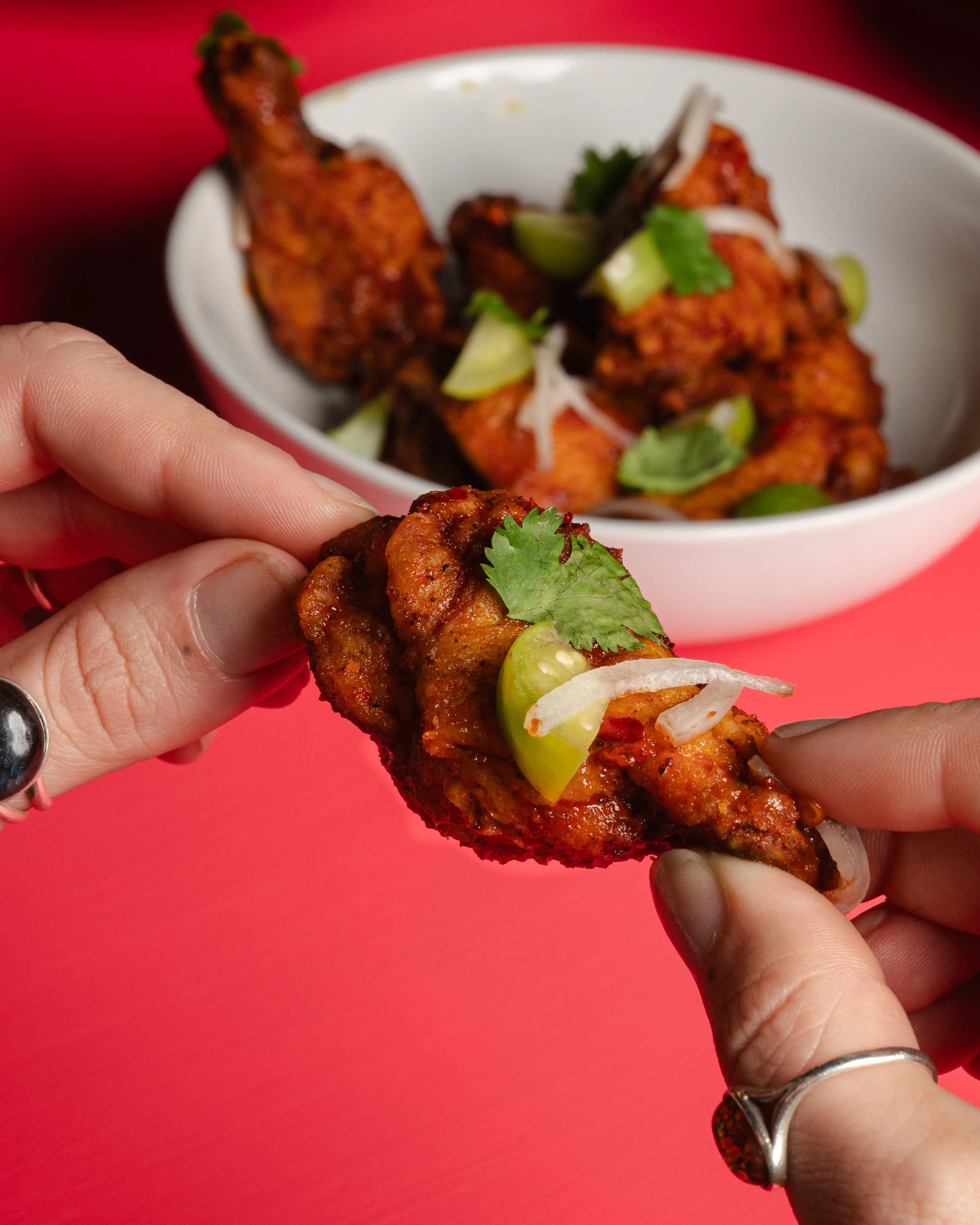 Person holding a piece of spicy chicken wing garnished with cilantro, onion, and a lime slice over a red background, with a bowl of more chicken wings in the background.