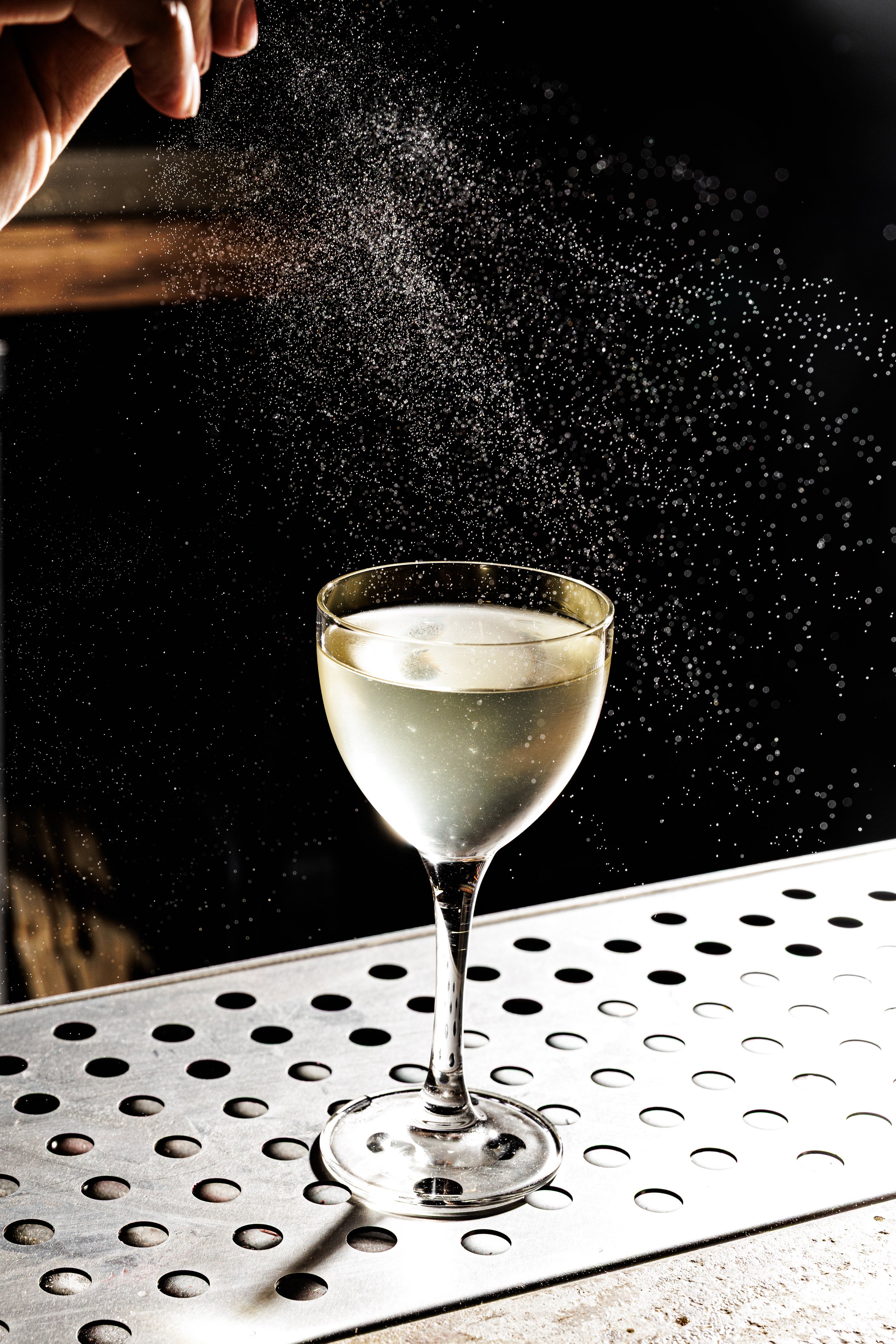A hand sprinkling powdered sugar into a glass of white wine on a bar counter.