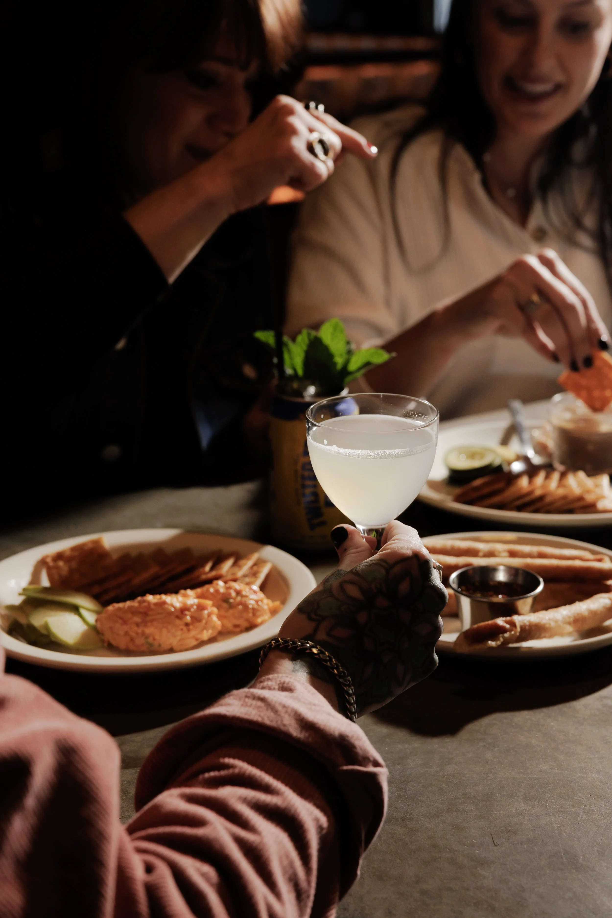 People enjoying drinks and food at a table in a dimly lit setting, with a hand holding a cocktail glass in focus.
