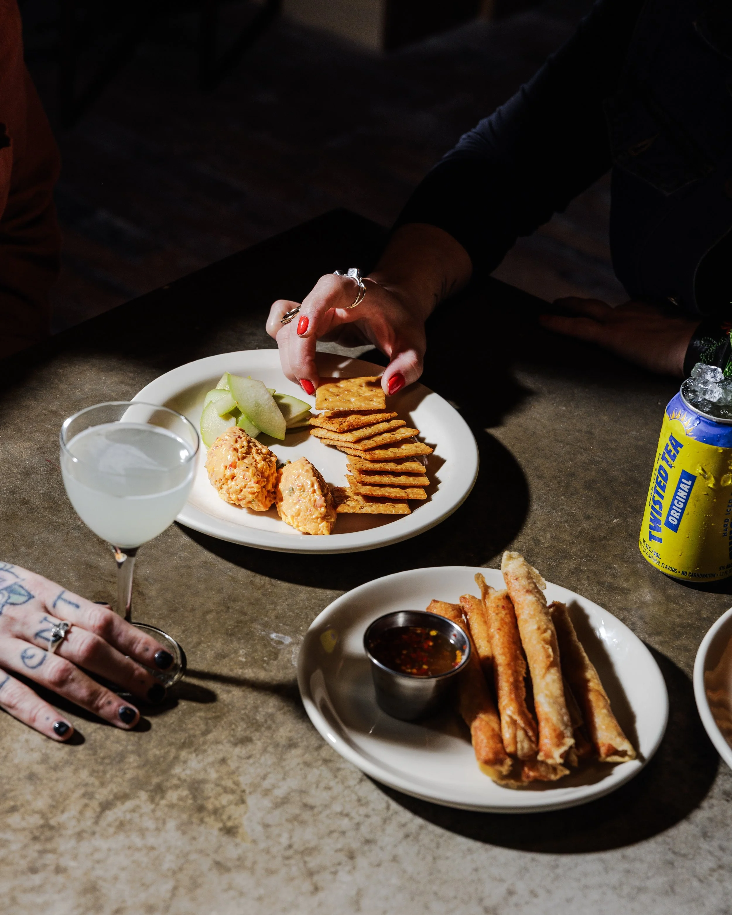 A person with manicured nails and rings reaches for crispy crackers on a white plate, which also has sliced melons and cheese balls. On the table, there is a glass of a pale drink, a plate of fried spring rolls with dipping sauce, and a can of Minute