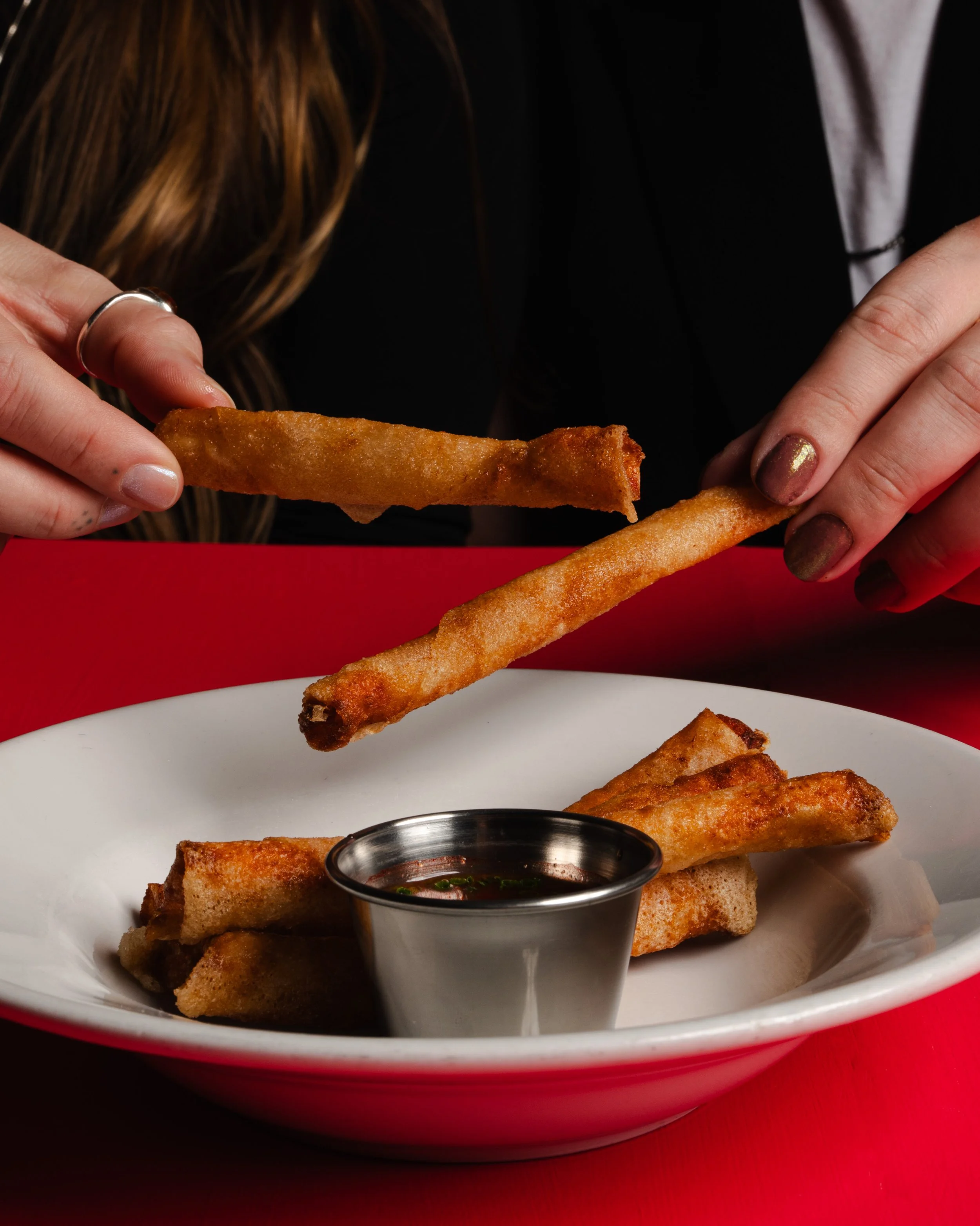 Person dipping fried spring roll into dipping sauce on a white plate with more spring rolls, red table, and black background