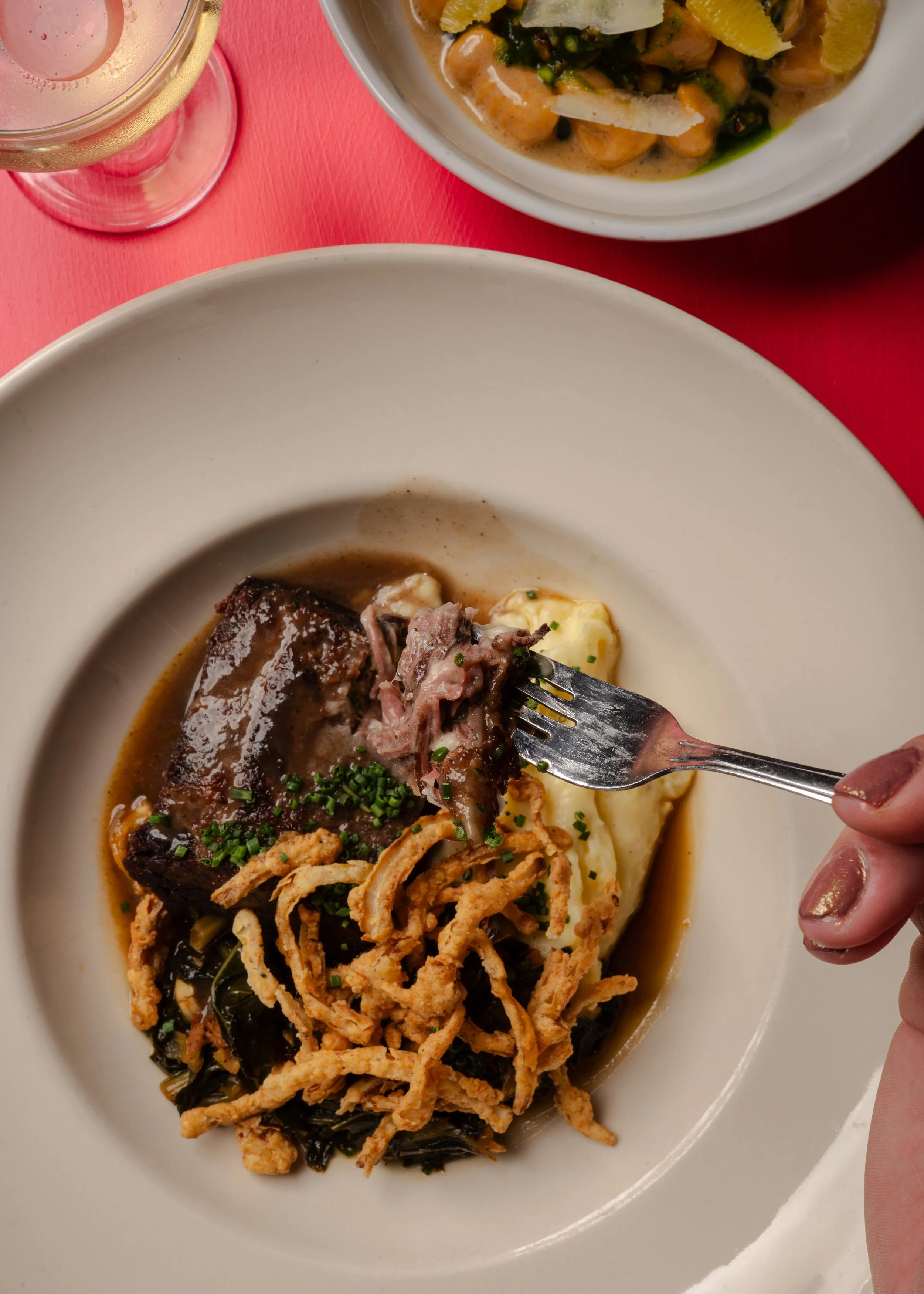 Close-up of a white plate with a serving of beef short ribs, mashed potatoes, and crispy onion strings, with a fork holding a piece of meat. In the background, there is a bowl of soup with vegetables and shrimp, and a glass of white wine on a red tab