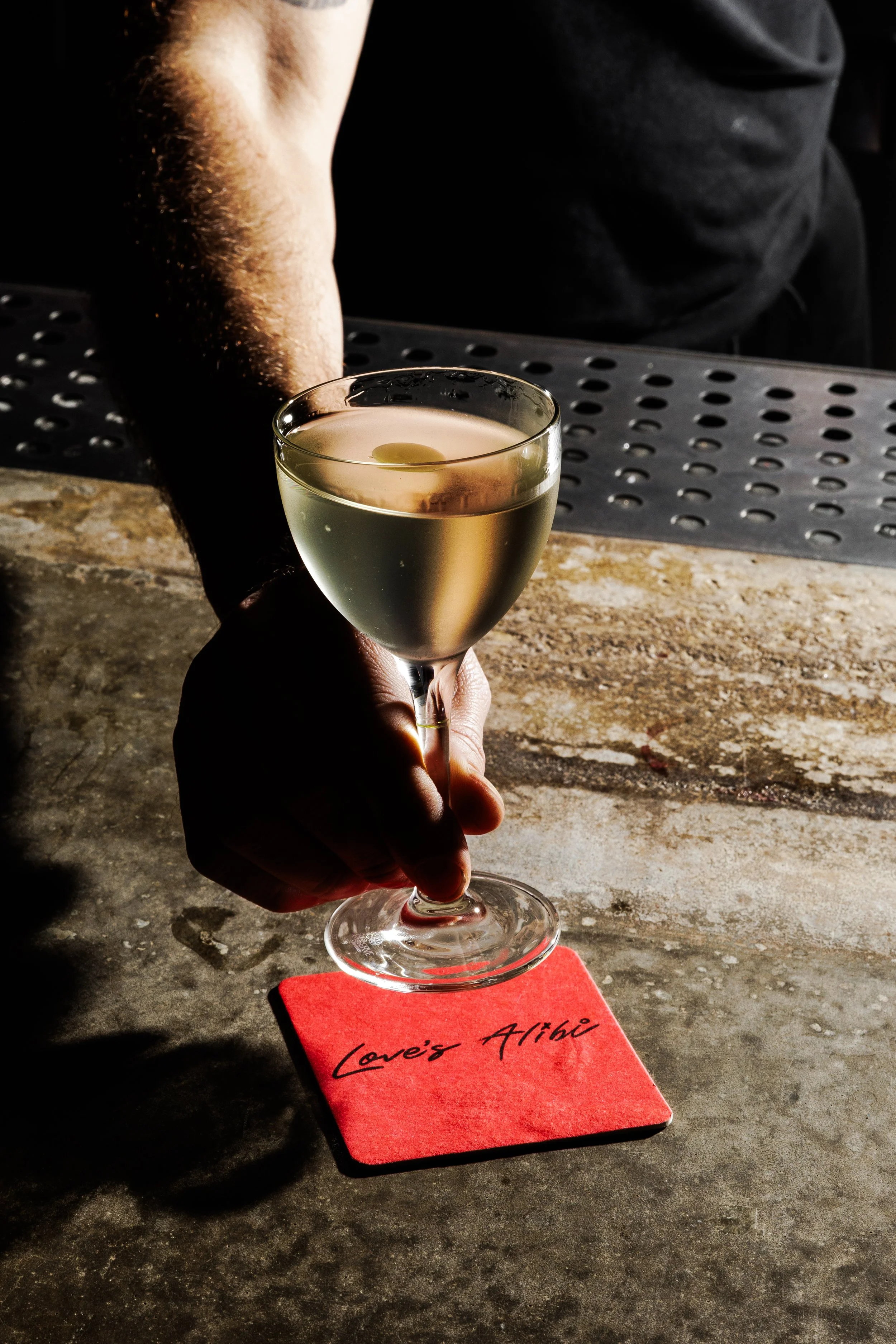 A person holding a cocktail glass with a light-colored drink, resting on a red napkin that says 'Love's Attic', on a textured bar counter.