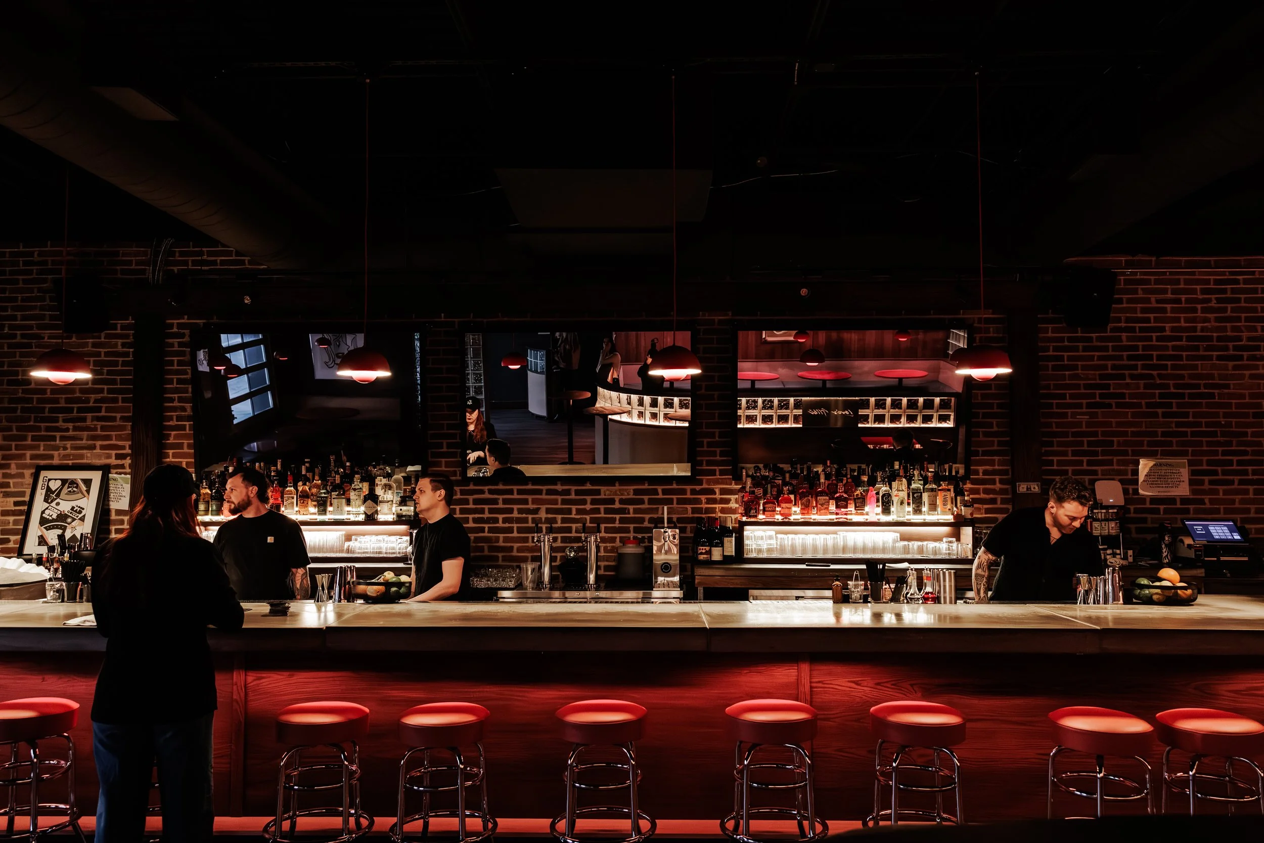 A dimly lit bar with a long marble counter, red bar stools, and a brick wall background. Several bartenders are working behind the counter, and a customer is standing in front of it, engaging with one of the bartenders. There are mirrors behind the b