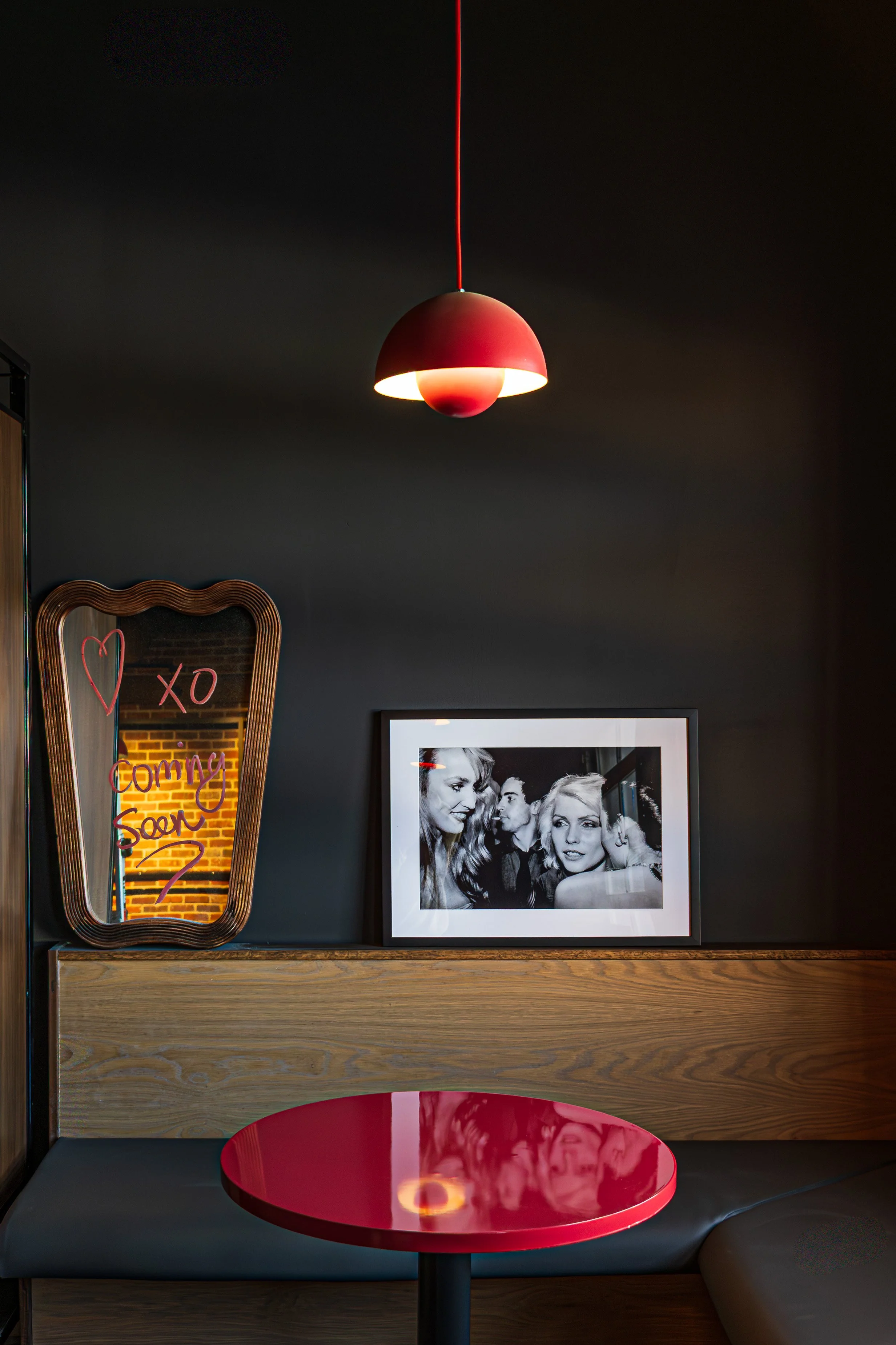 A cozy corner inside a stylish cafe featuring a red round table, a black and white framed photo of four cheerful people, a wavy-edged mirror with a neon sign reading 'Xo Coming Soon,' and a black wall with a red pendant light hanging from the ceiling