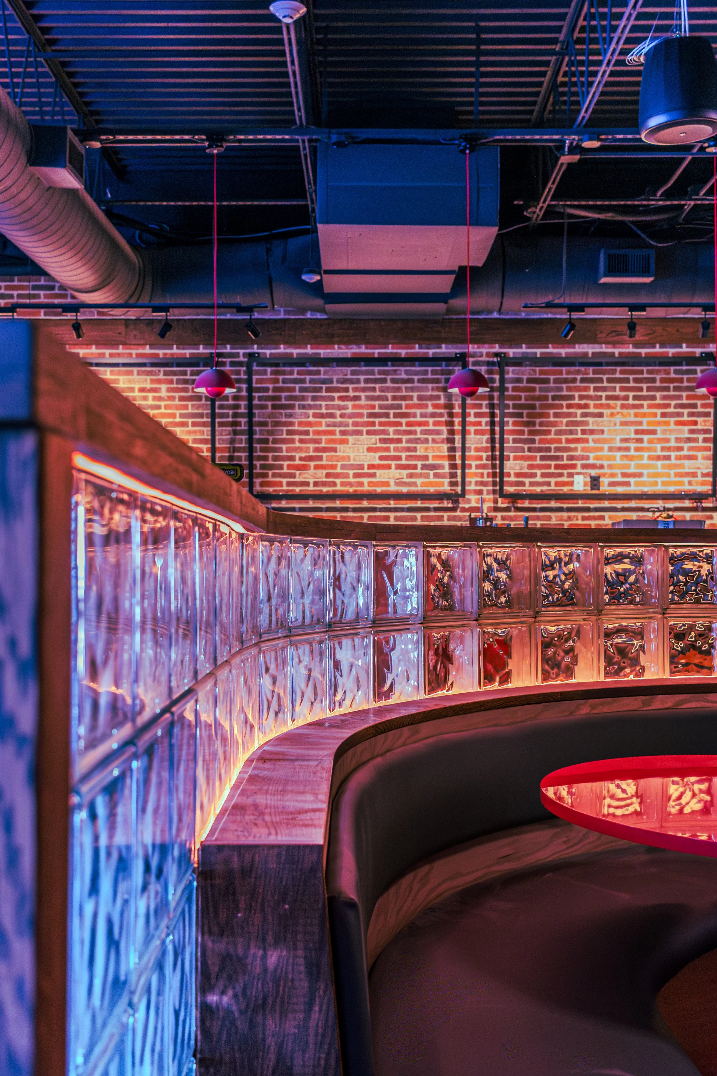 Interior of a modern bar or restaurant with exposed brick walls, glass block railings illuminated with colorful lights, pendant lighting, and visible ventilation ducts and ceiling pipes.