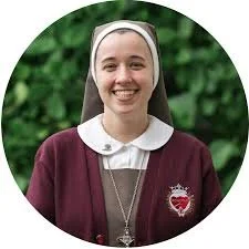 A nun, smiling, with a white headband and a badge, outdoors with green foliage in the background.