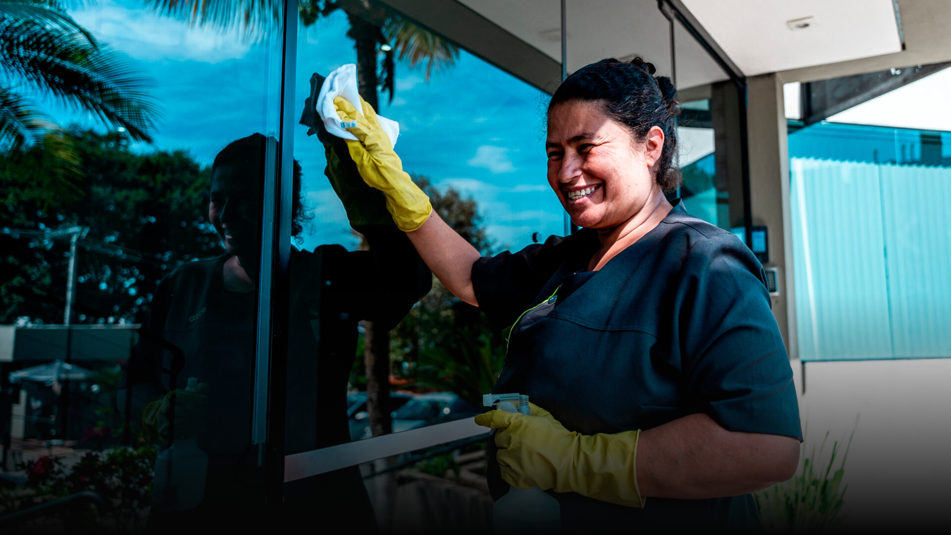 Mulher sorridente limpando uma janela com pano, usando luvas amarelas, ao ar livre