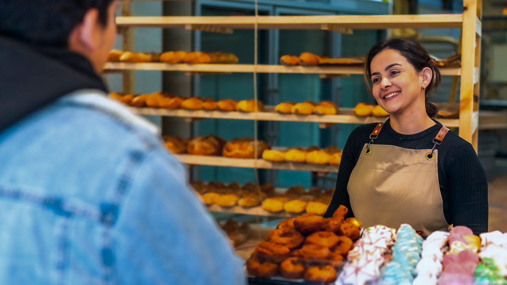 Vendedora sorridente atendendo cliente em banca de doces, com diversas opções de bolos e doces coloridos na frente e prateleiras com mais doces ao fundo.