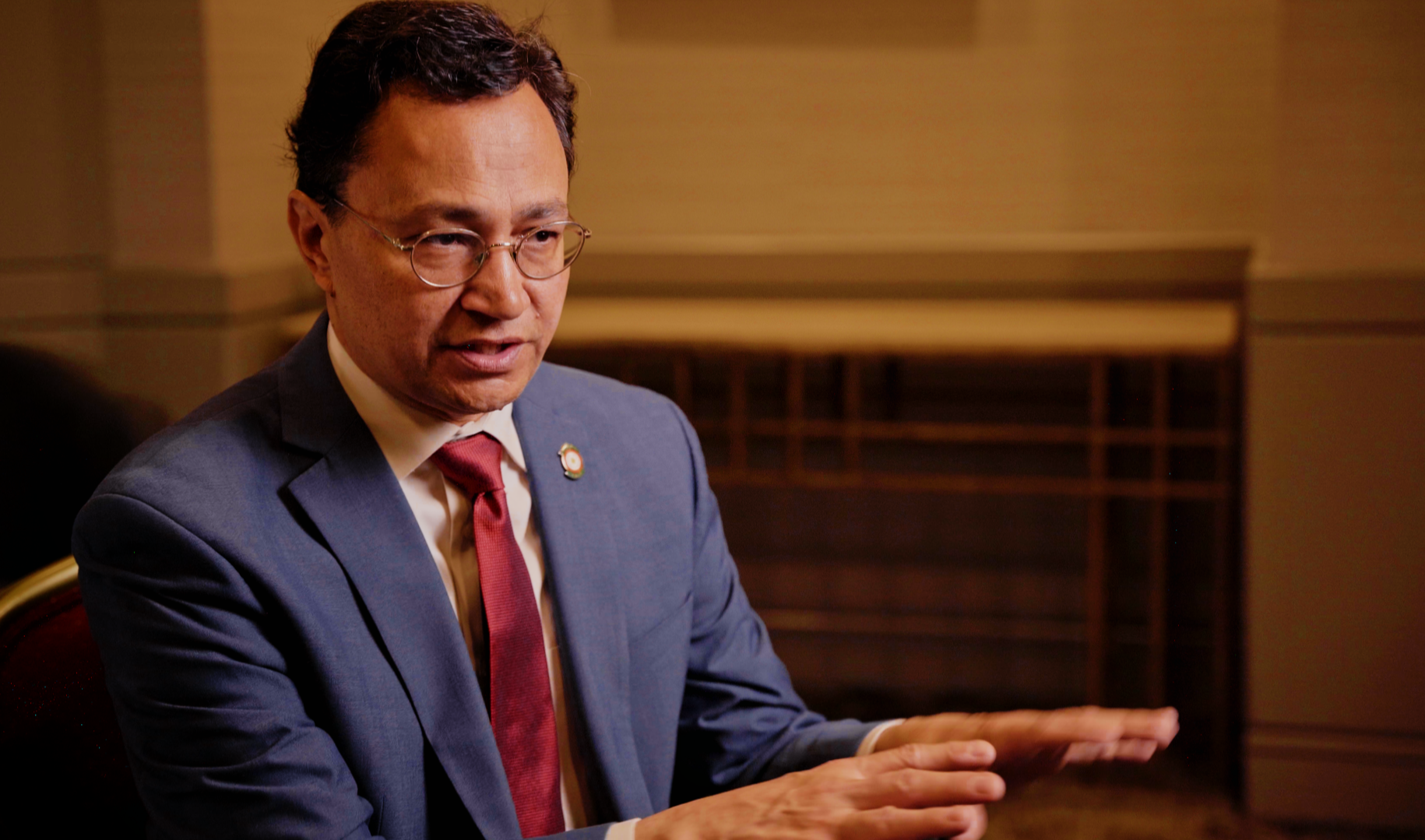 A man in a suit and tie sitting at a table, gesturing with his hands during a conversation or interview.