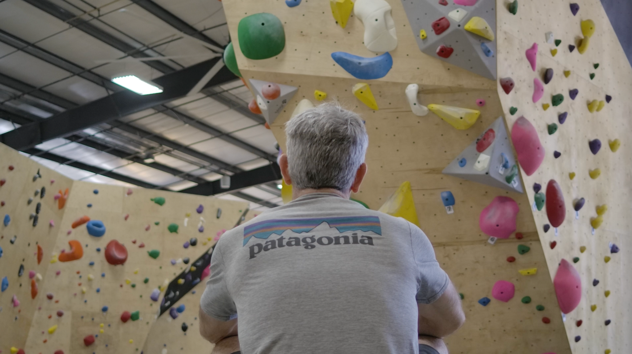 Back of an older man with gray hair sitting and looking at an indoor climbing gym wall with colorful holds.
