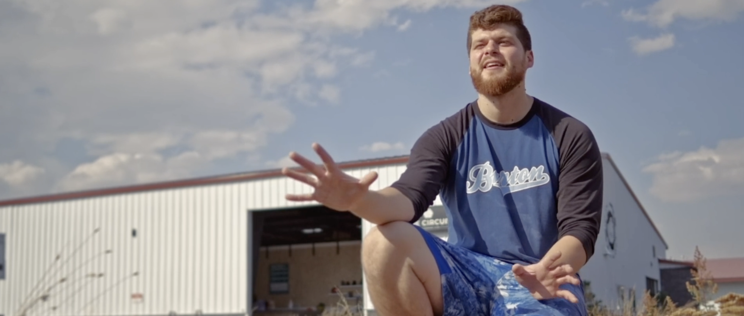 Man with a beard and short brown hair sitting outside on a sunny day, wearing a blue and black long-sleeve shirt and blue shorts, gesturing with his hand. There is a large industrial-style building with a white exterior and red roof in the background