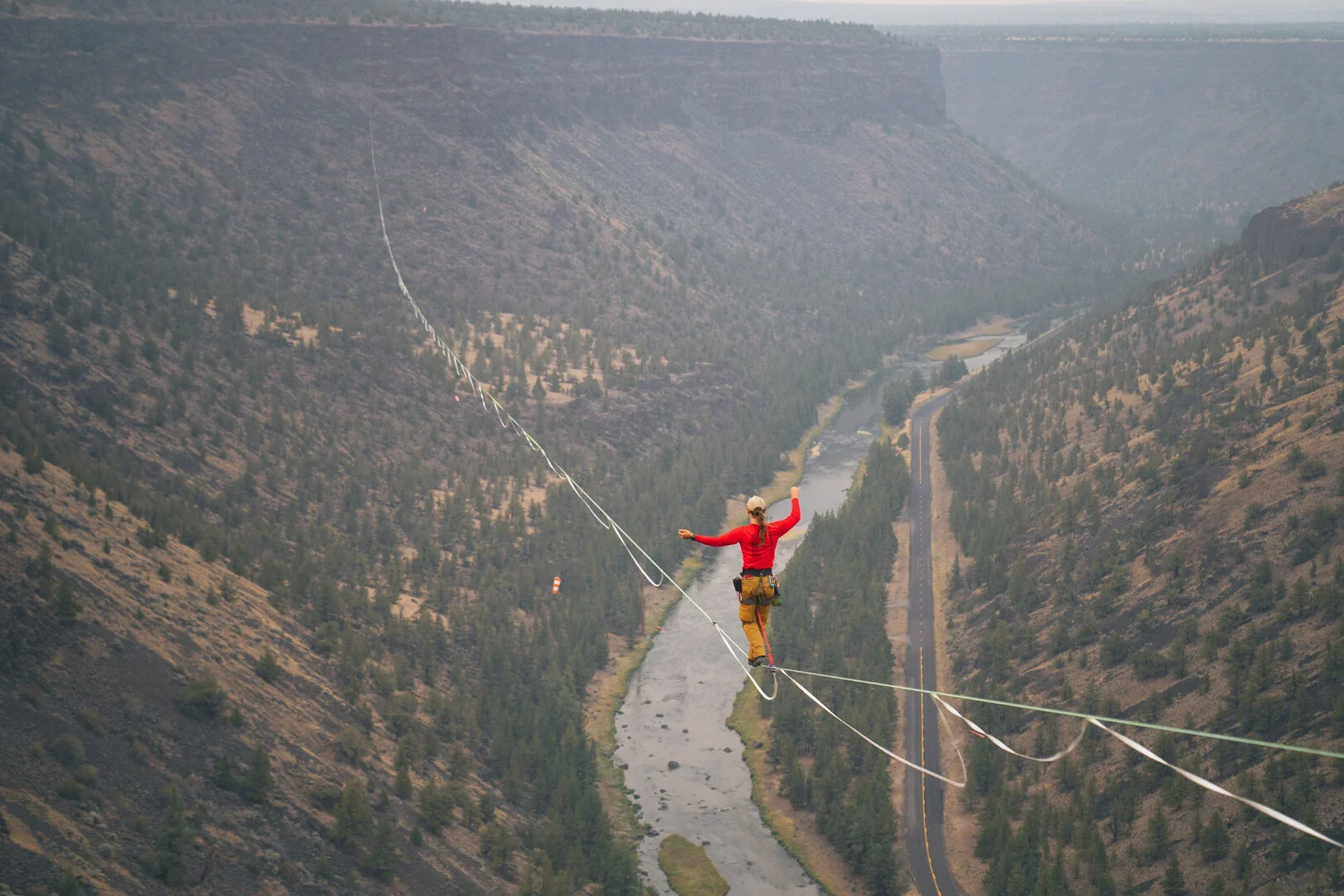 A tightrope walker balancing on a high wire stretched across a canyon with a river and trees below, wearing a red top and tan pants.