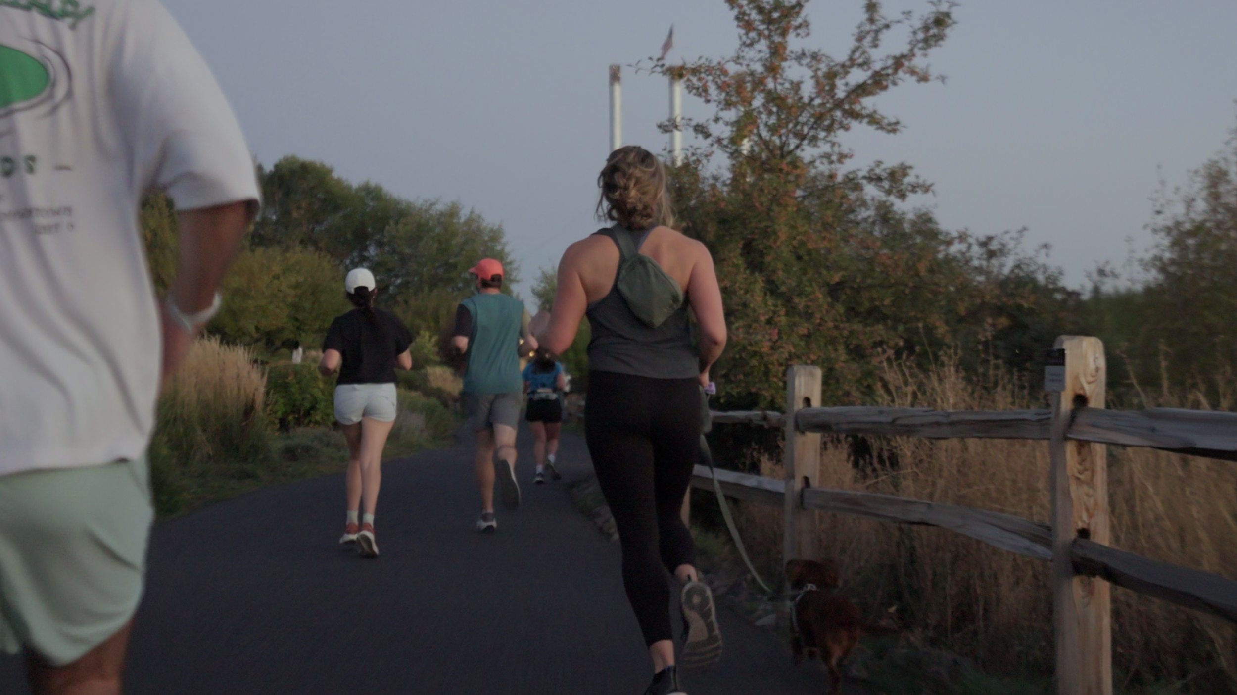 People running on a paved trail next to a wooden fence on a scenic outdoor trail during sunset.