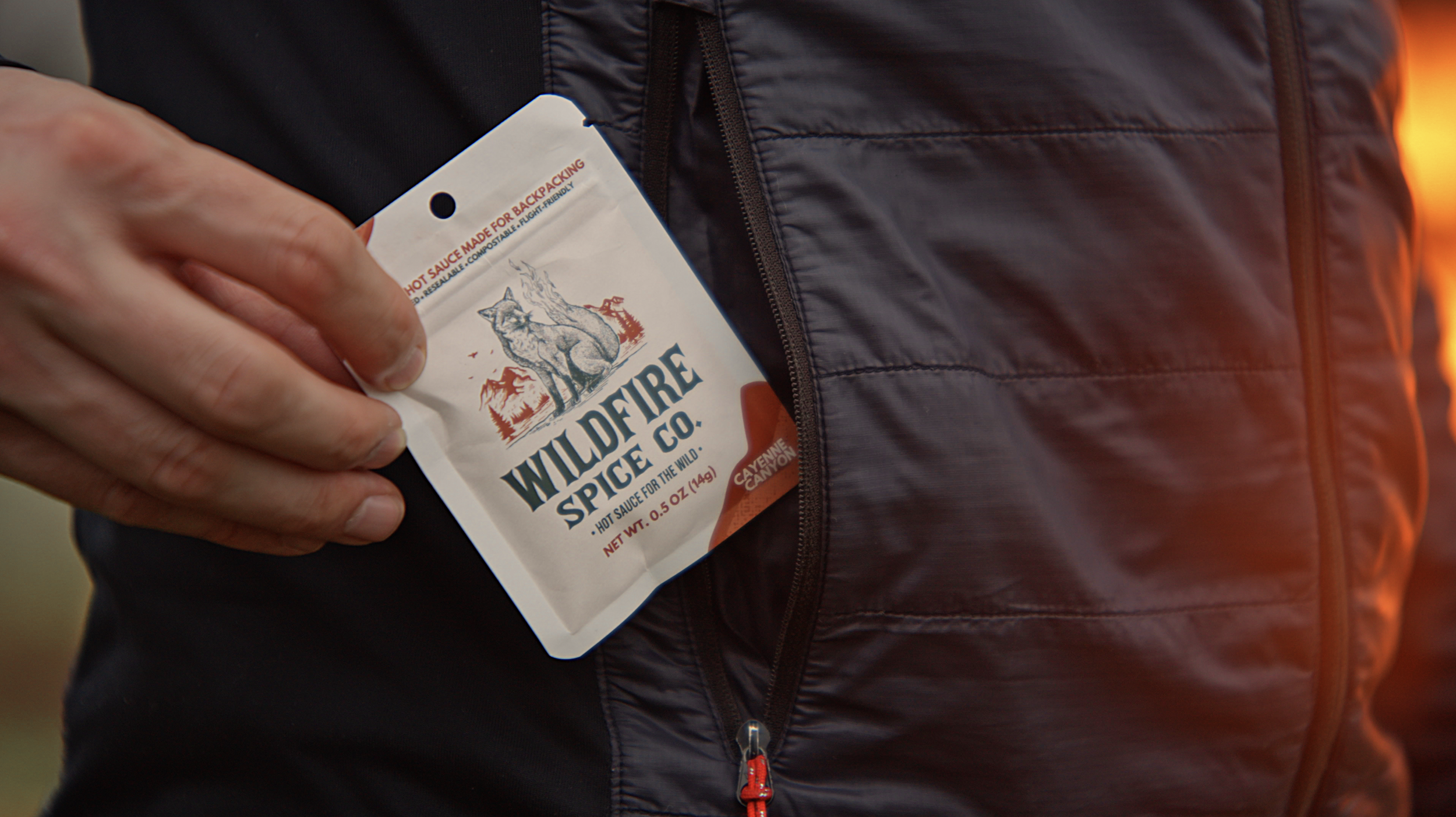 Person holding a Wilderness Spice Co. hot sauce packet, standing outdoors near a black puffy jacket.