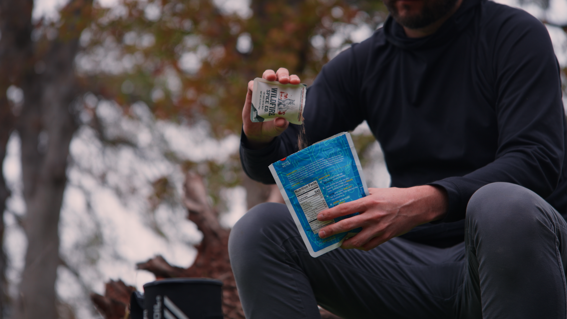 A person wearing a black long-sleeve shirt and gray pants sitting outdoors with trees in the background, pouring seasoning from a spice packet onto a food item in a blue plastic container.