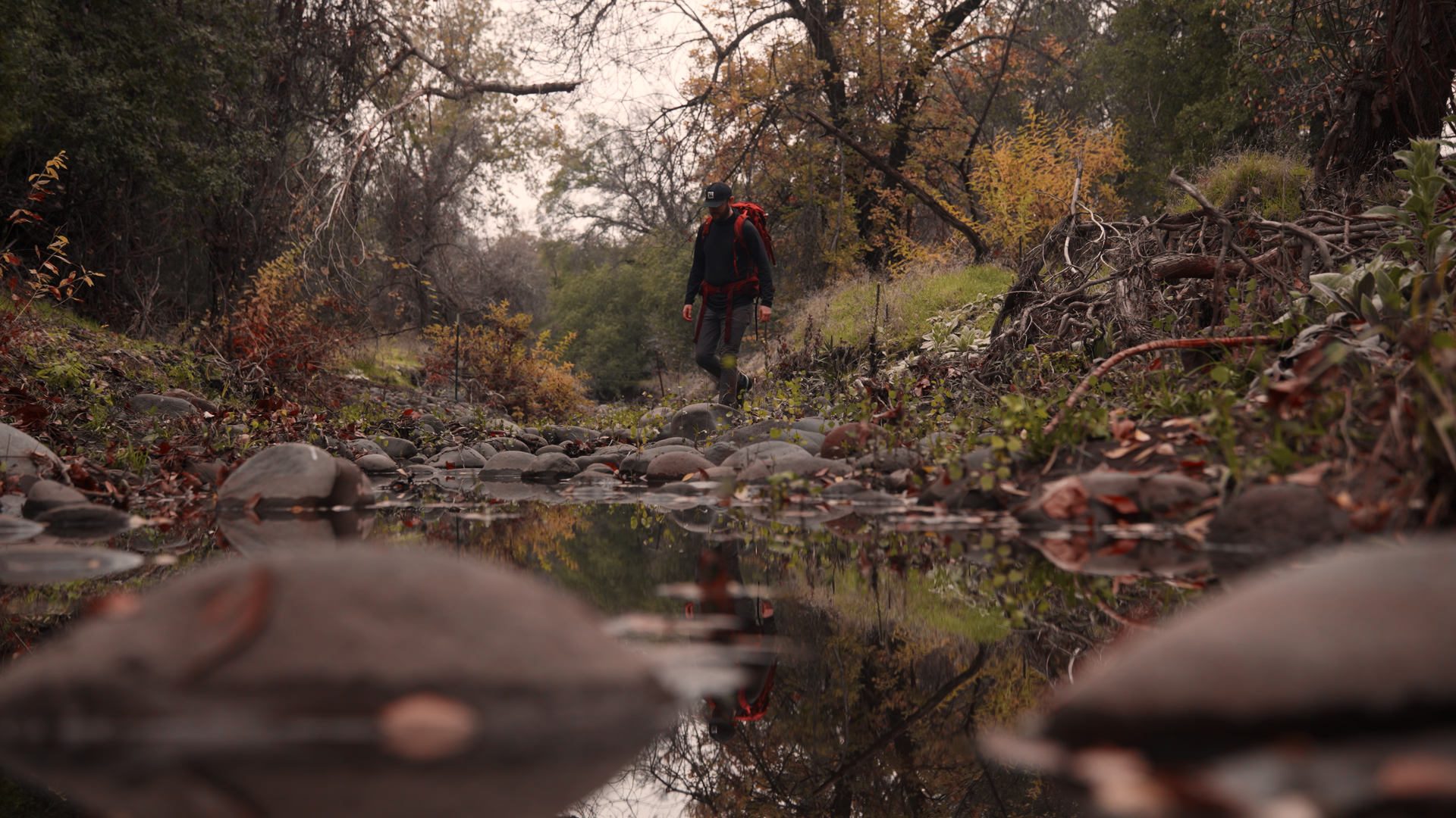 A person hiking in a forested area during fall, walking along a rocky creek with reflections of trees on the water, wearing a black outfit with a red backpack and cap.