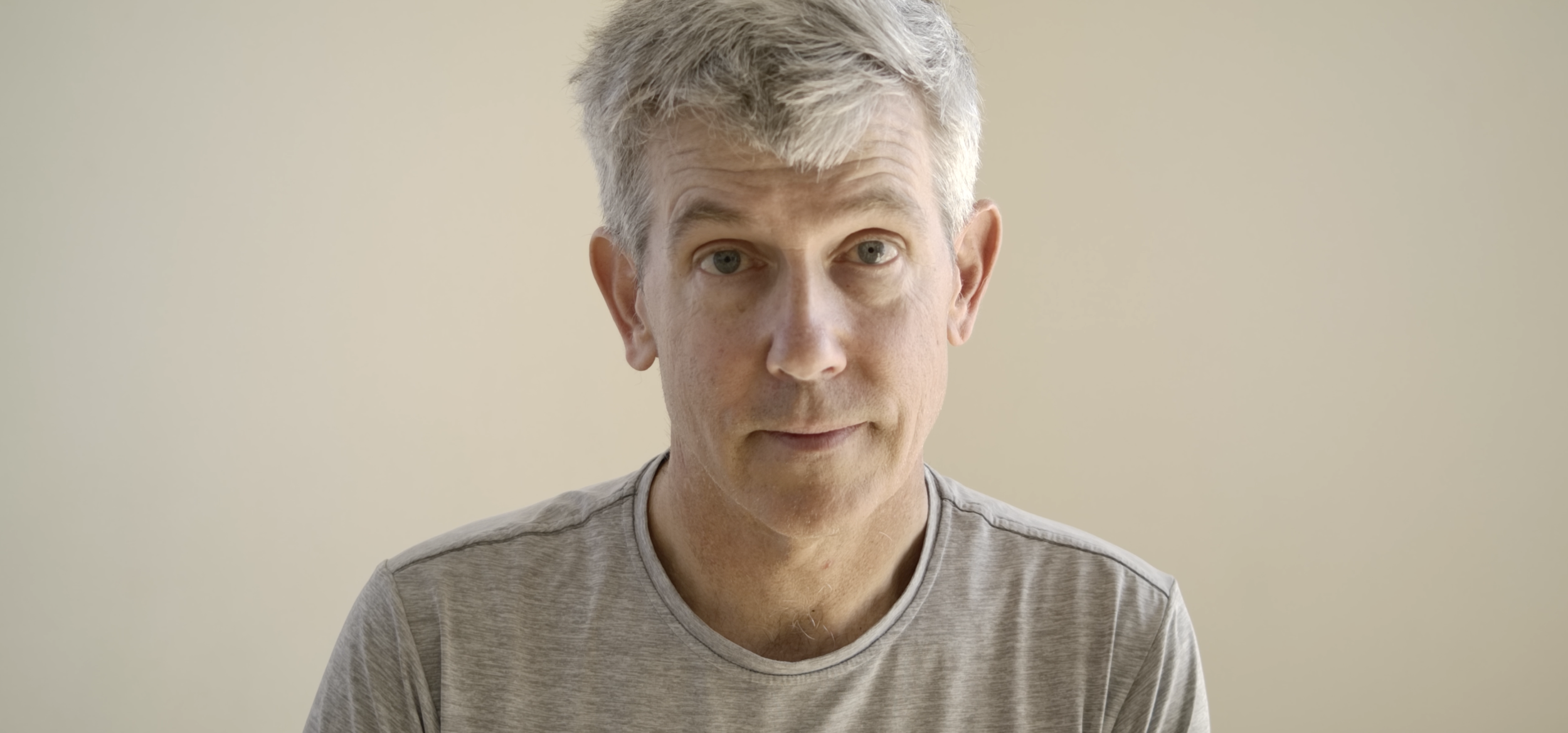 Close-up of a middle-aged man with gray hair and blue eyes wearing a light gray t-shirt, looking directly at the camera with a neutral expression, against a beige background.