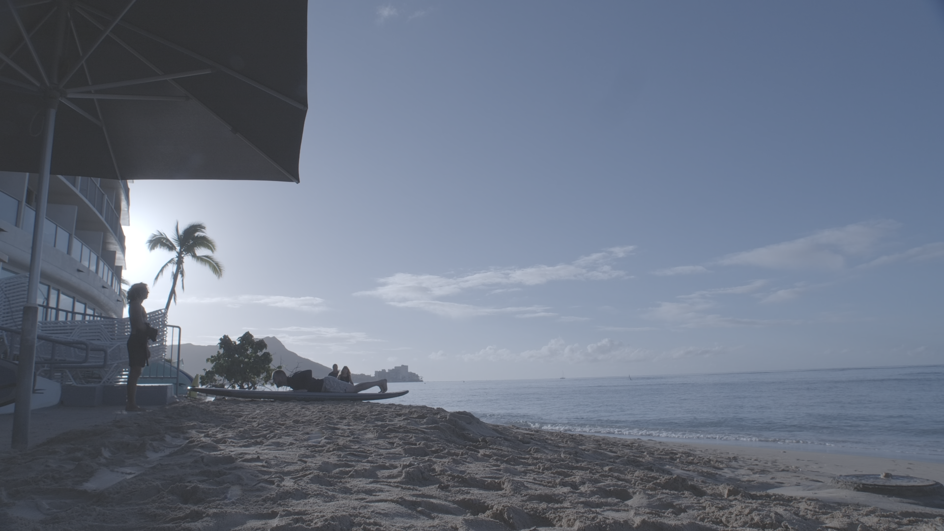 Beach scene with a person lying on a surfboard and others sitting near the shoreline, palm trees, a building, and mountains in the background under a partly cloudy sky.