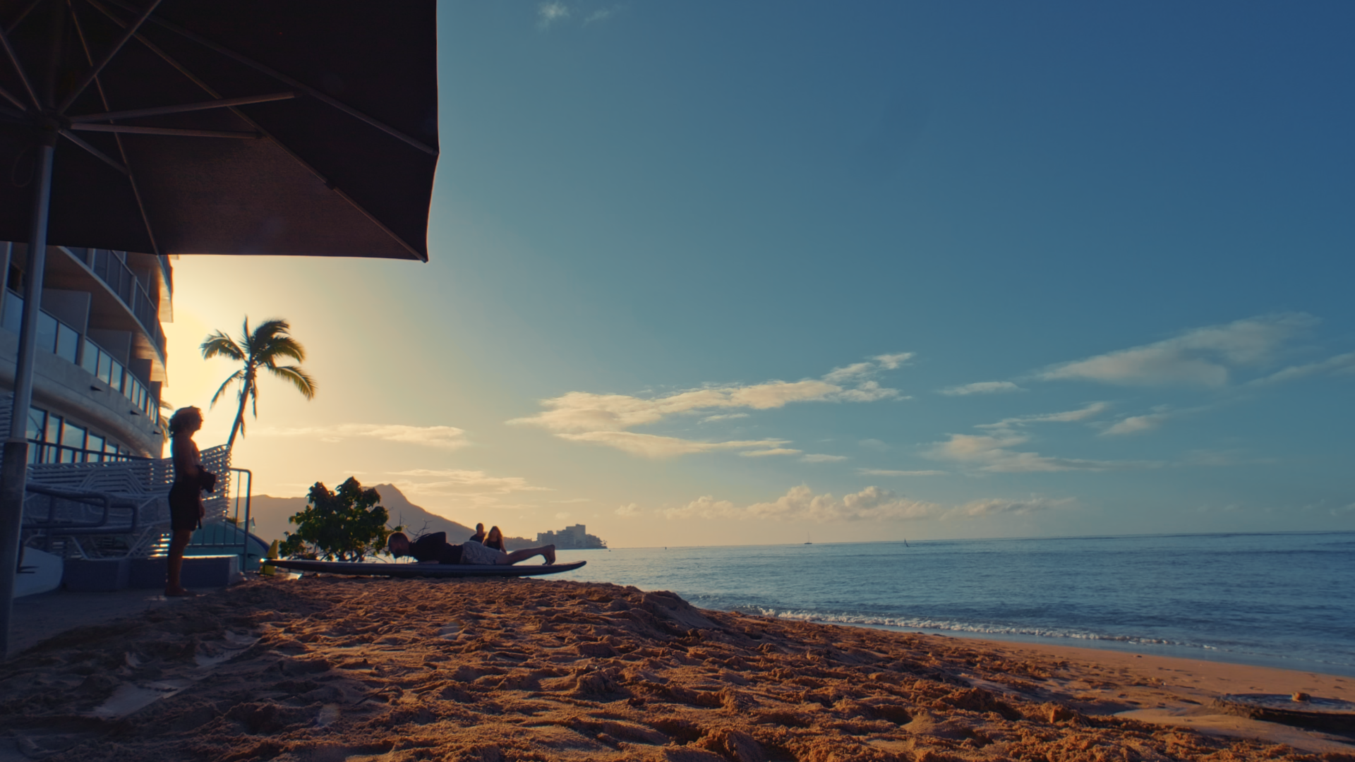 Silhouettes of people on a sandy beach at sunset, with a person standing and two lying on surfboards, near palm trees and a building, with a mountain in the background and a calm ocean.