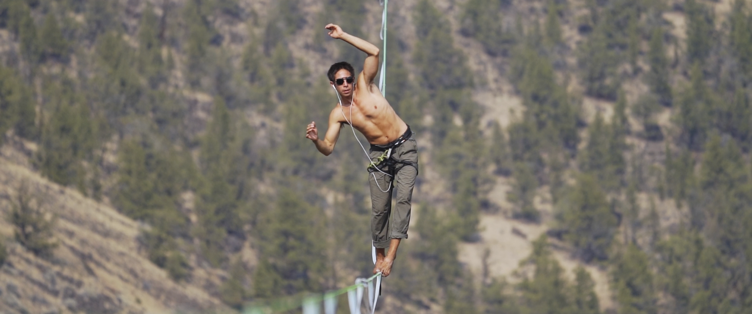 A shirtless man with sunglasses walking a slackline outdoors with a forested mountain in the background.