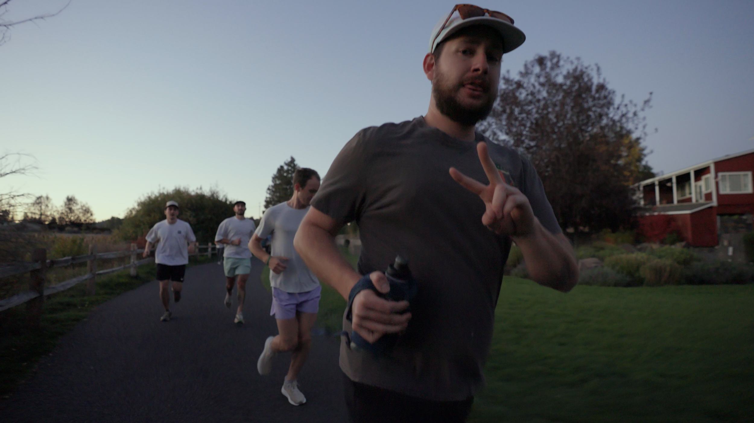Man in running attire making a peace sign while jogging on a paved path with three other runners in the background, in an outdoor park during dusk.
