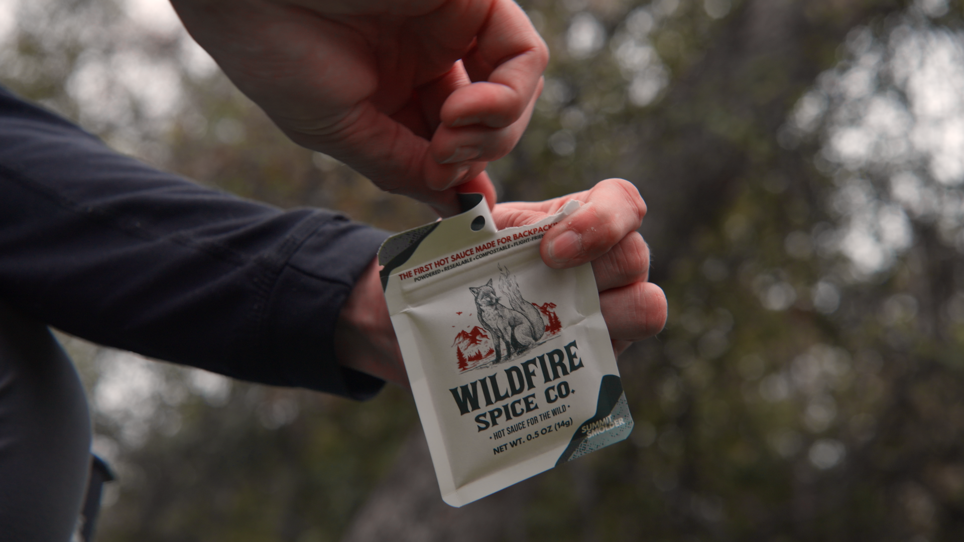 A person opening a packet of Wilderness Spice Co. hot sauce for backpacking, featuring a wolf and mountain illustrations on the packaging, outdoors with blurred trees in the background.
